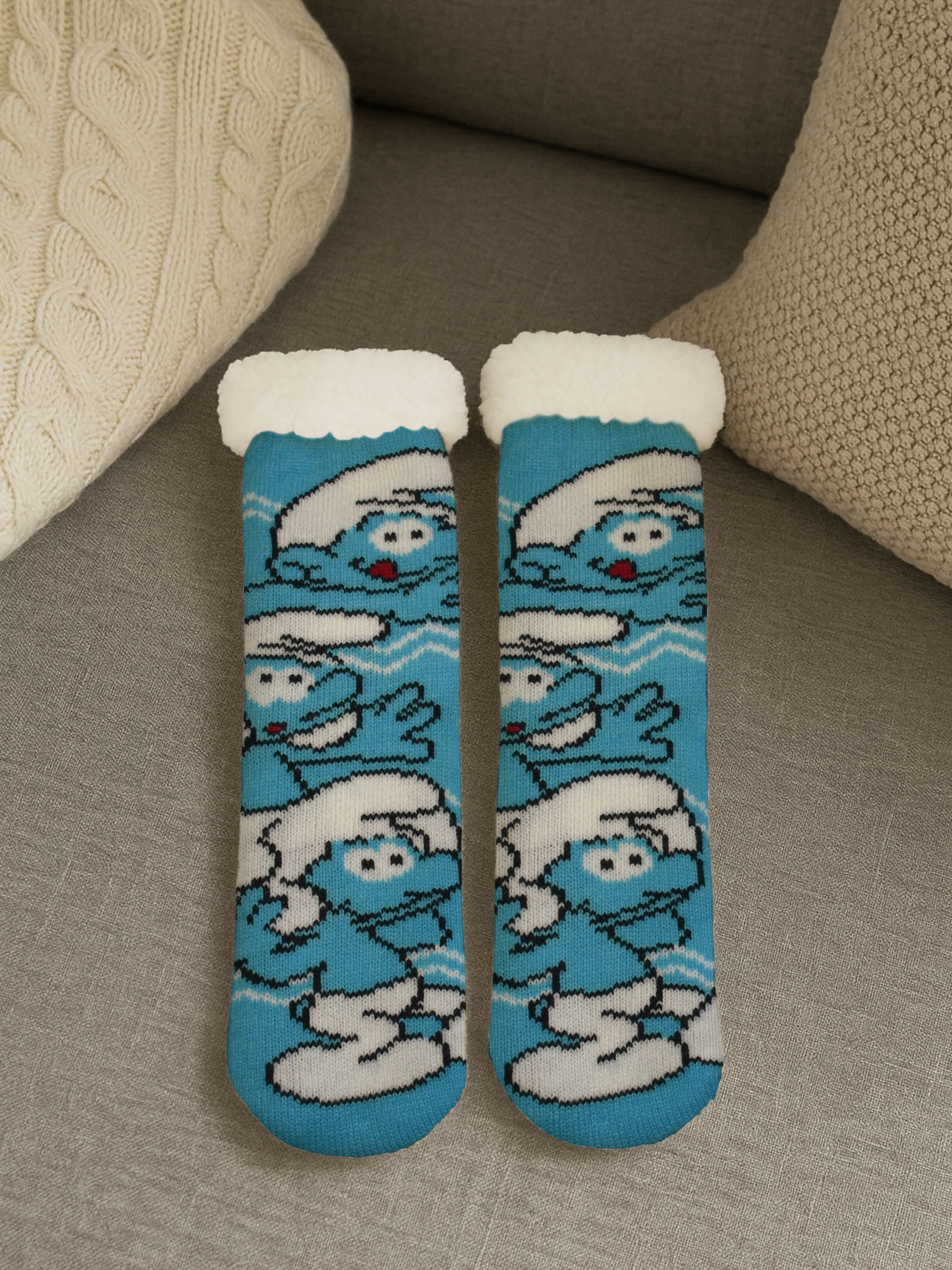Zwei blaue Schlumpfsocken mit weißer Verzierung und Schlumpf-Charakteren liegen auf einer grauen Oberfläche, mit Kissen im Hintergrund.