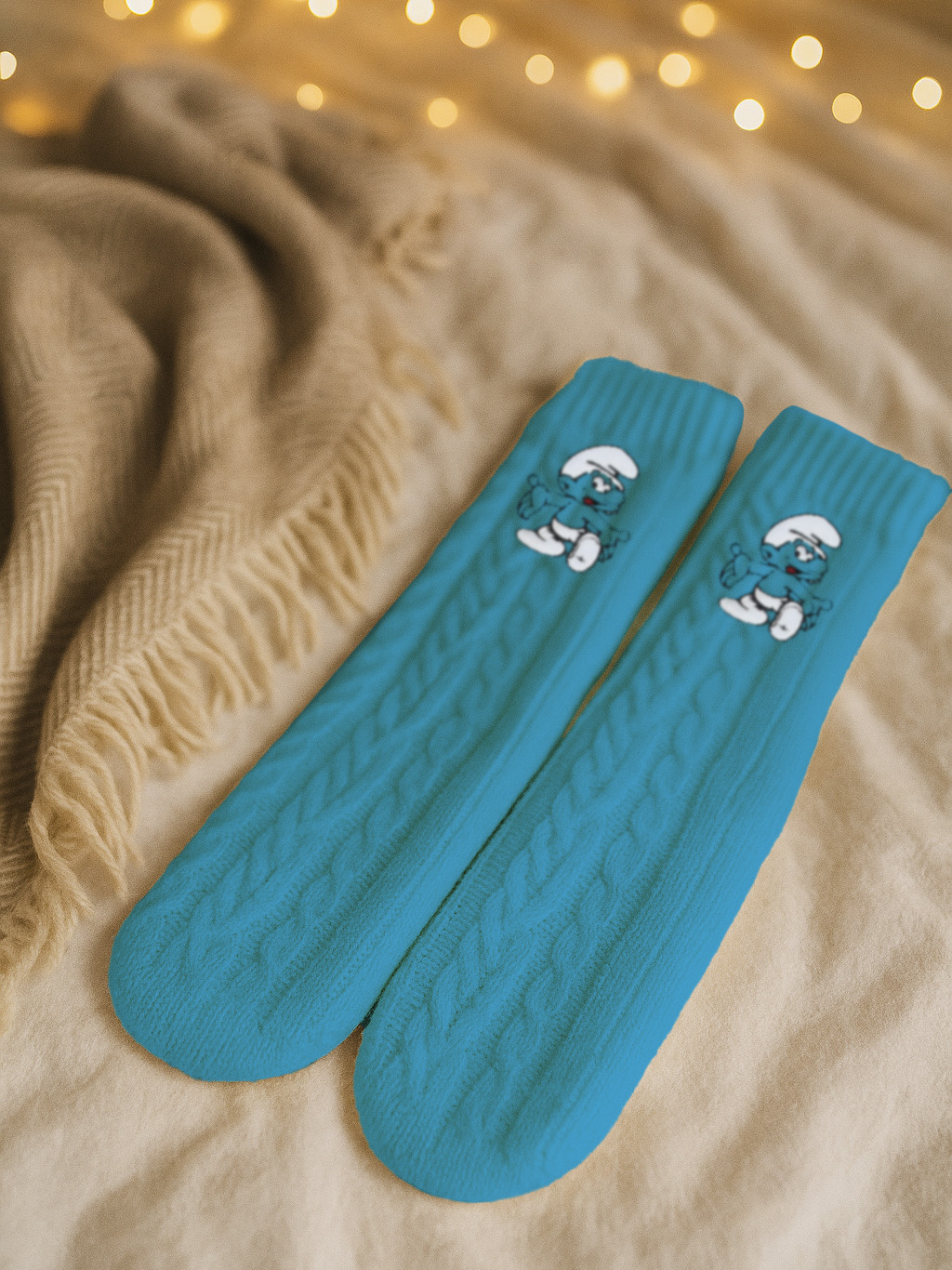 Zwei blaue Schlumpf-Socken mit Zopfmuster, liegen auf einer beigen Decke. Ein beiger Schal und unscharfe Lichter im Hintergrund.