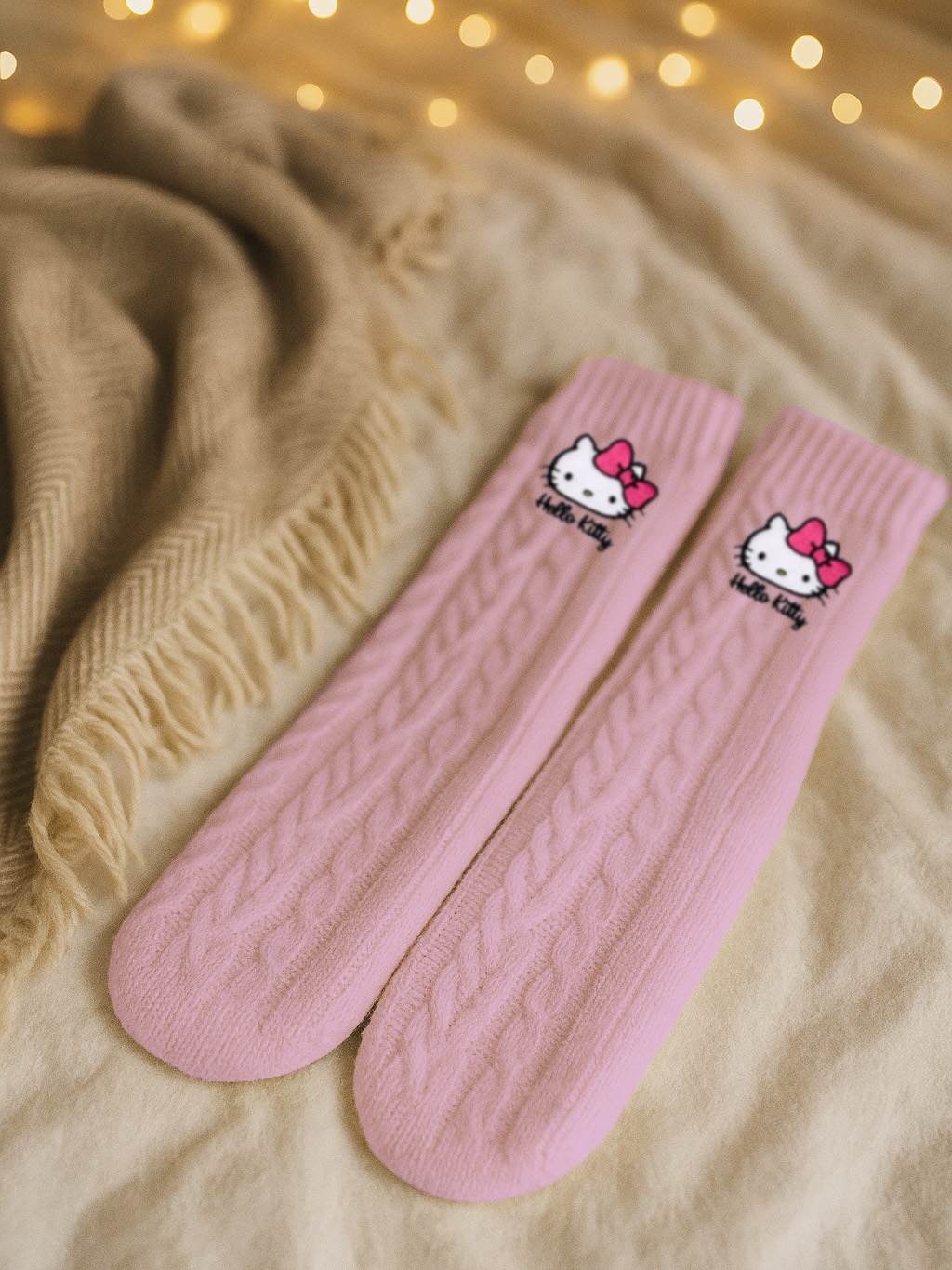 Zwei rosa Hello Kitty Socken auf einem Bett. Eine beige Decke und Bokeh-Lichter im Hintergrund.