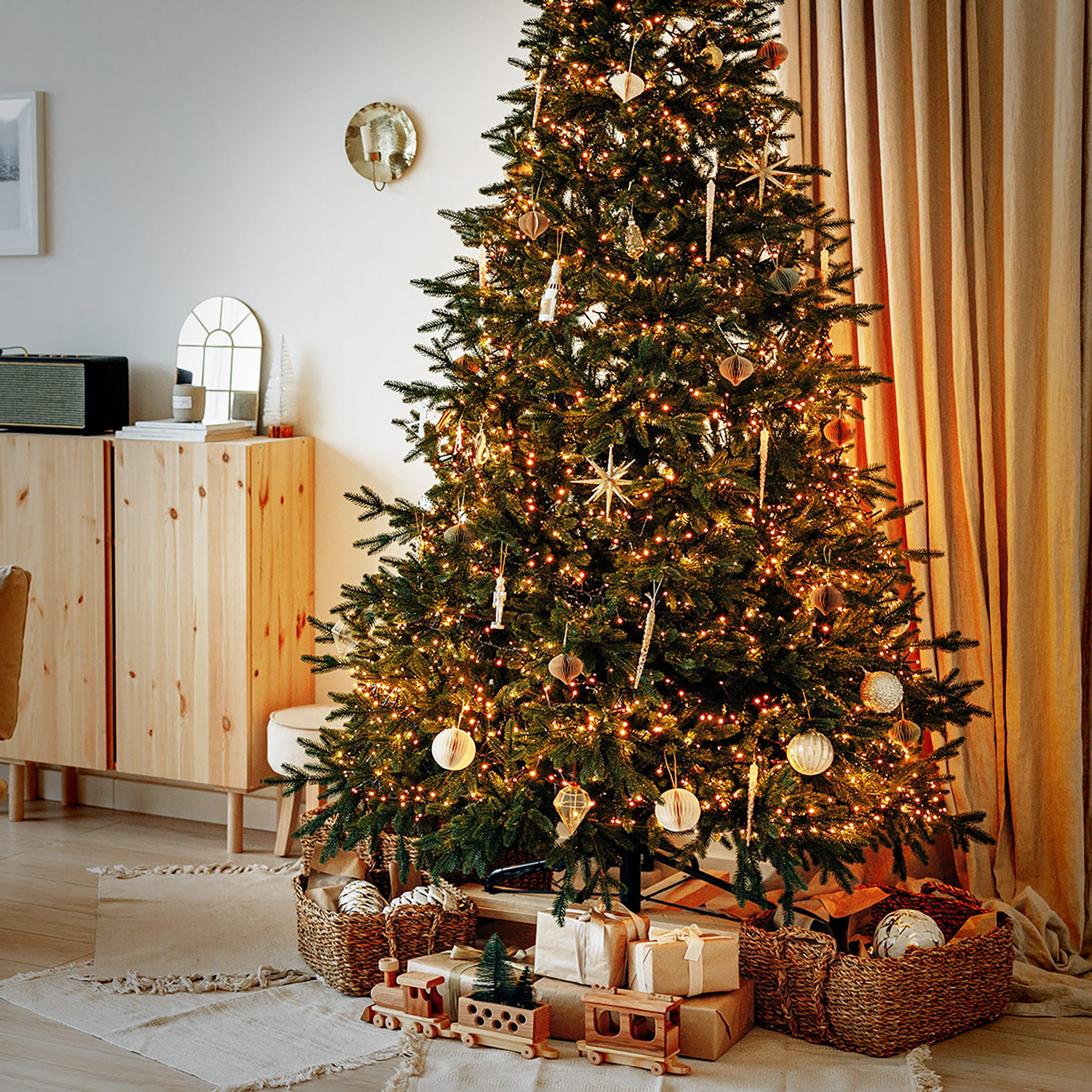 Ein geschmückter Weihnachtsbaum mit Lichtern und Ornamenten. Geschenke und ein Spielzeugzug sind darunter platziert. Ein Raum in neutralen Tönen.