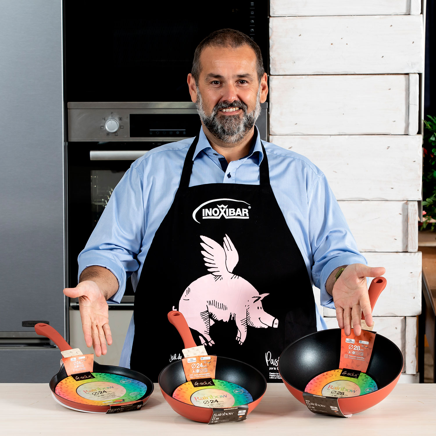 Un hombre con un delantal con un gráfico de cerdo presenta tres sartenes. Las sartenes están sobre una mesa. El fondo presenta una cocina.