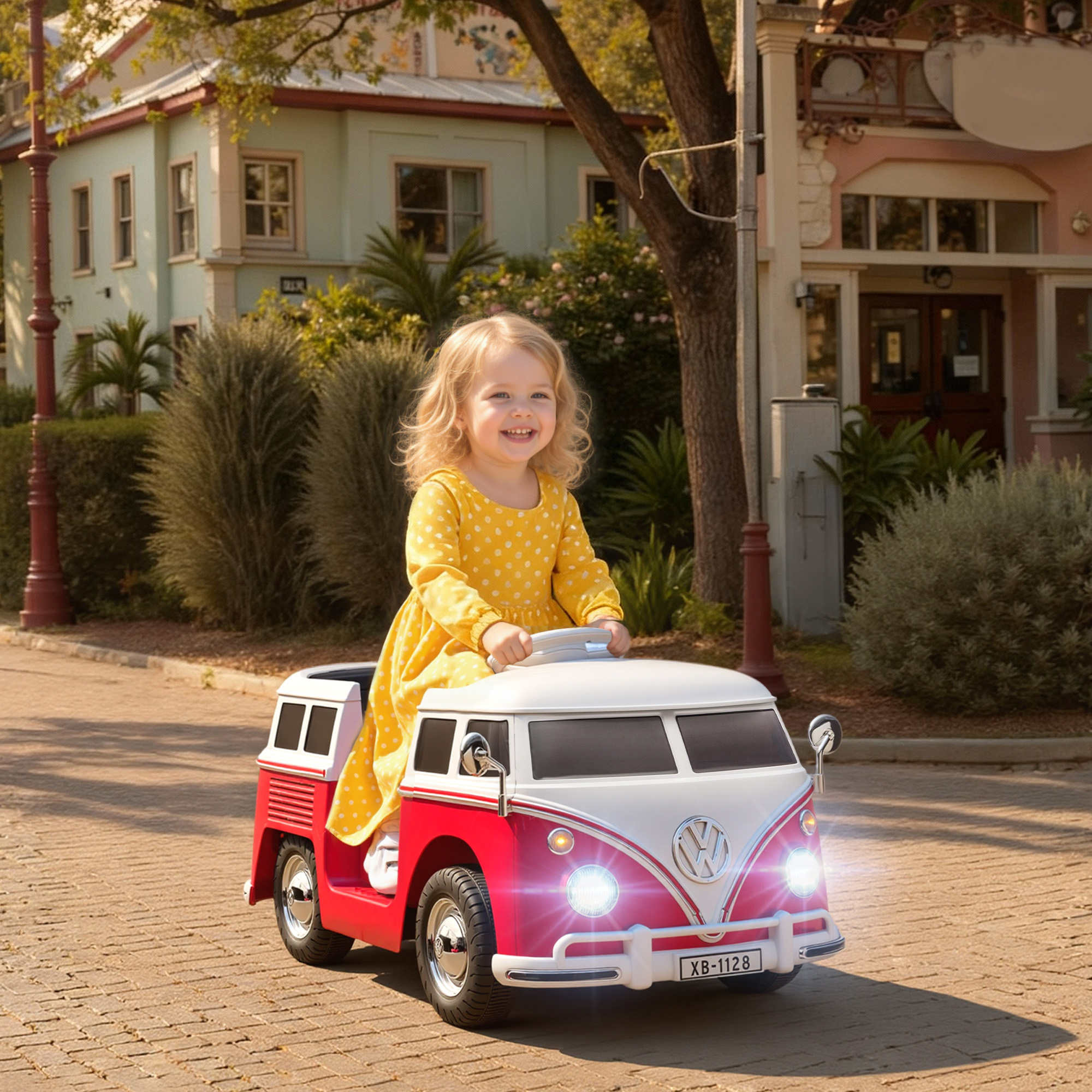 Un bambino sorridente con un vestito giallo guida un'auto giocattolo rossa e bianca su una strada di mattoni, alberi ed edifici sullo sfondo.