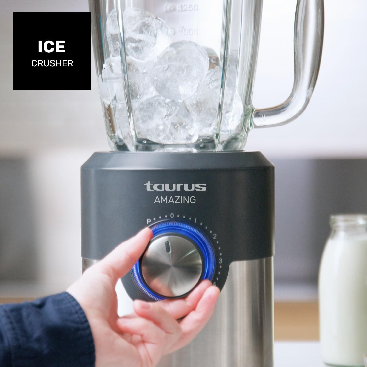 Una mano gira el dial de una licuadora llena de hielo. Un tarro de vidrio con leche se encuentra en el fondo. La licuadora dice 'Taurus Amazing'.