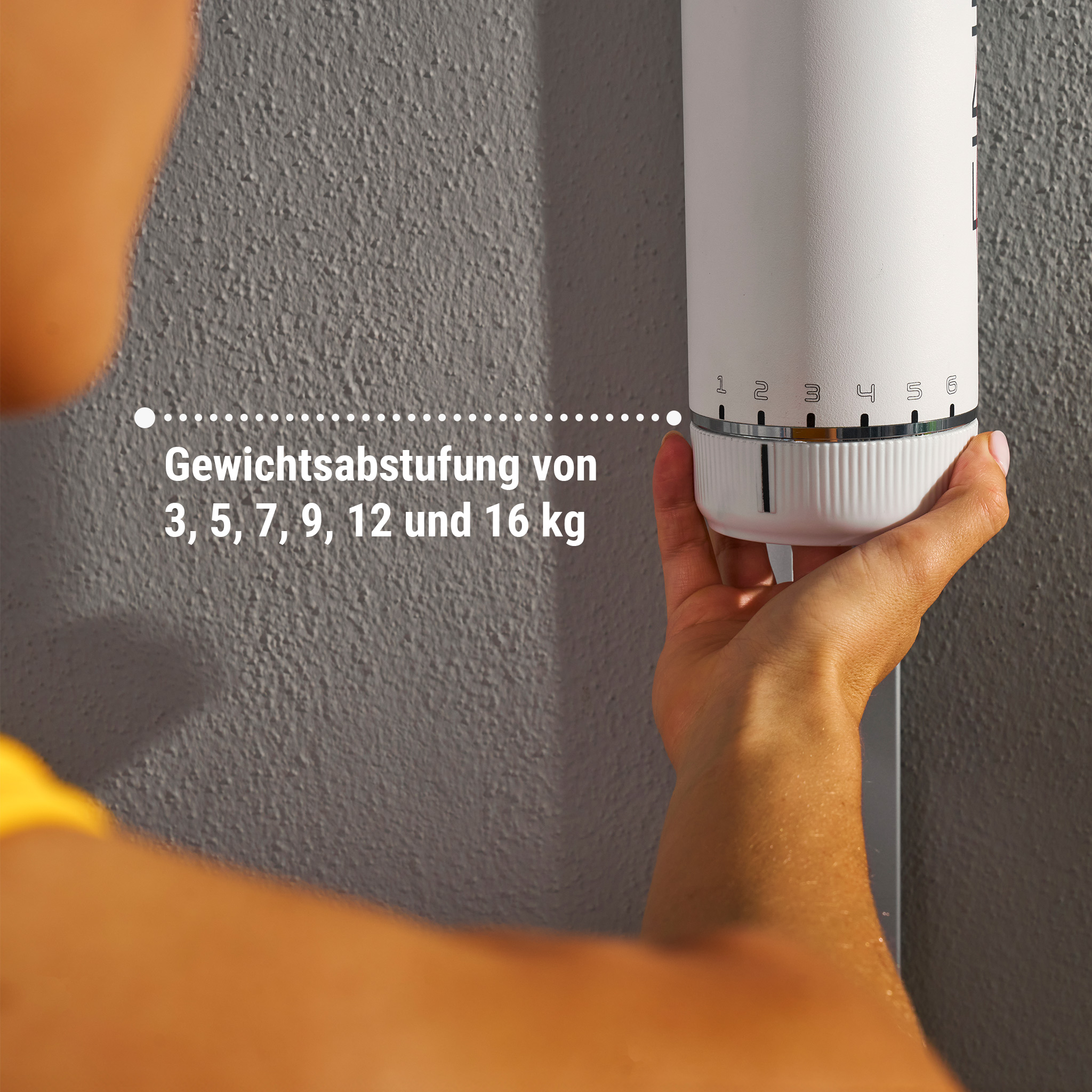 Eine Person stellt ein weißes Gewichtseinstellgerät an einer grauen Wand ein. Die Zahlen 1-6 sind sichtbar.
