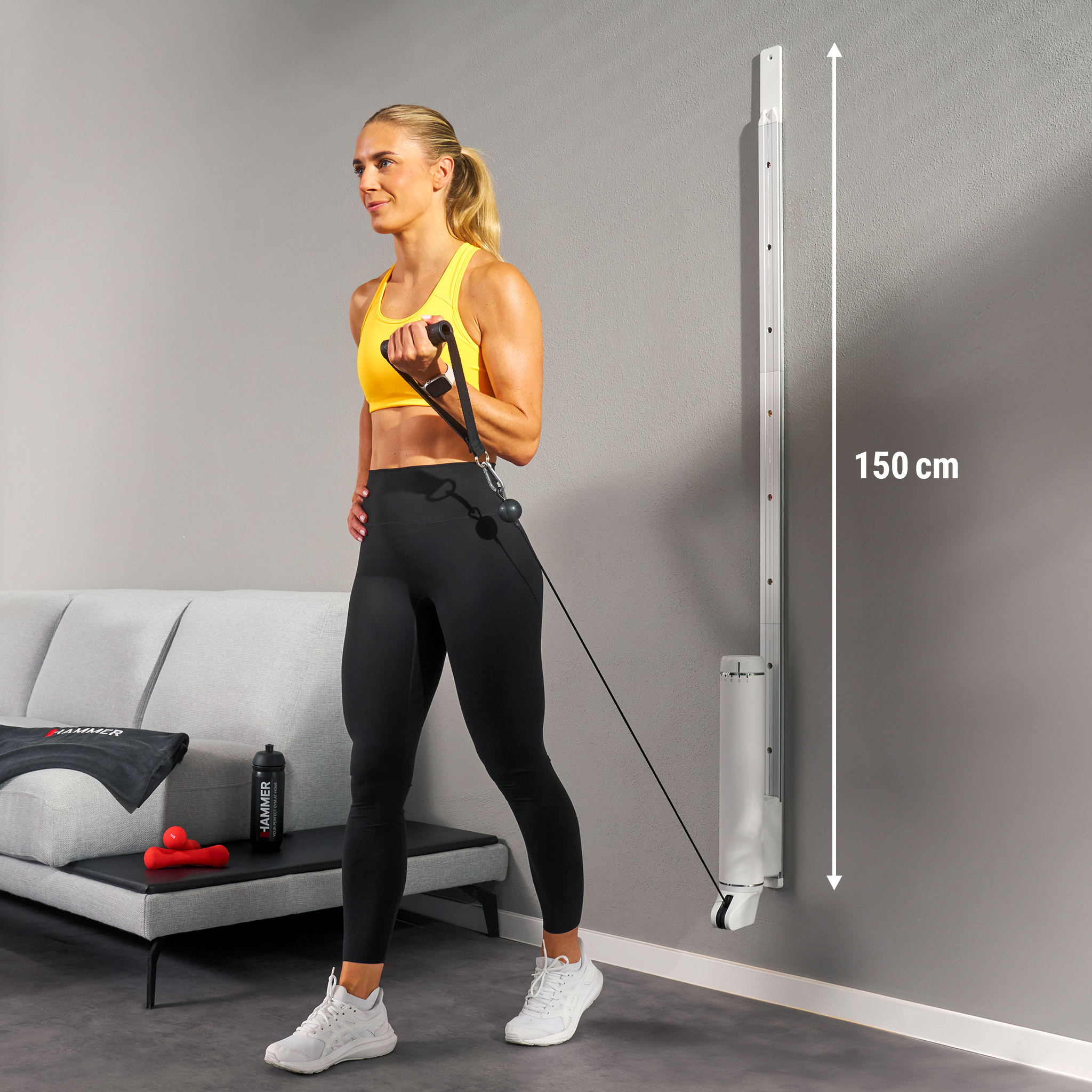 Frau, die mit einem Widerstandsband trainiert und ein an der Wand befestigtes Fitnessgerät benutzt. Im Hintergrund sind Sofa und Gewichte sichtbar.
