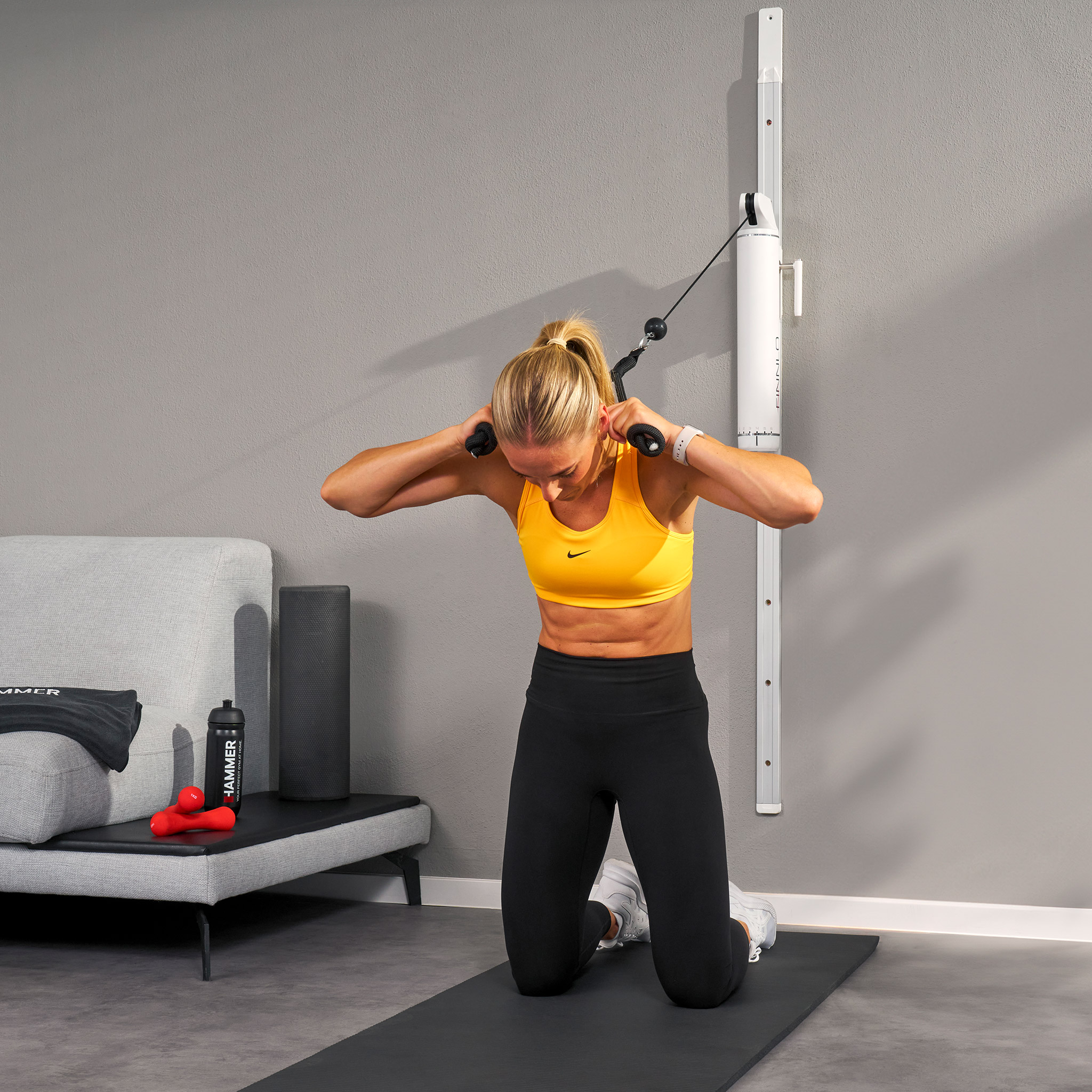 Frau kniet auf Matte und benutzt Kabelzug. Sie trägt gelbes Oberteil, schwarze Hose. Fitnessgeräte und Sofa im Hintergrund.
