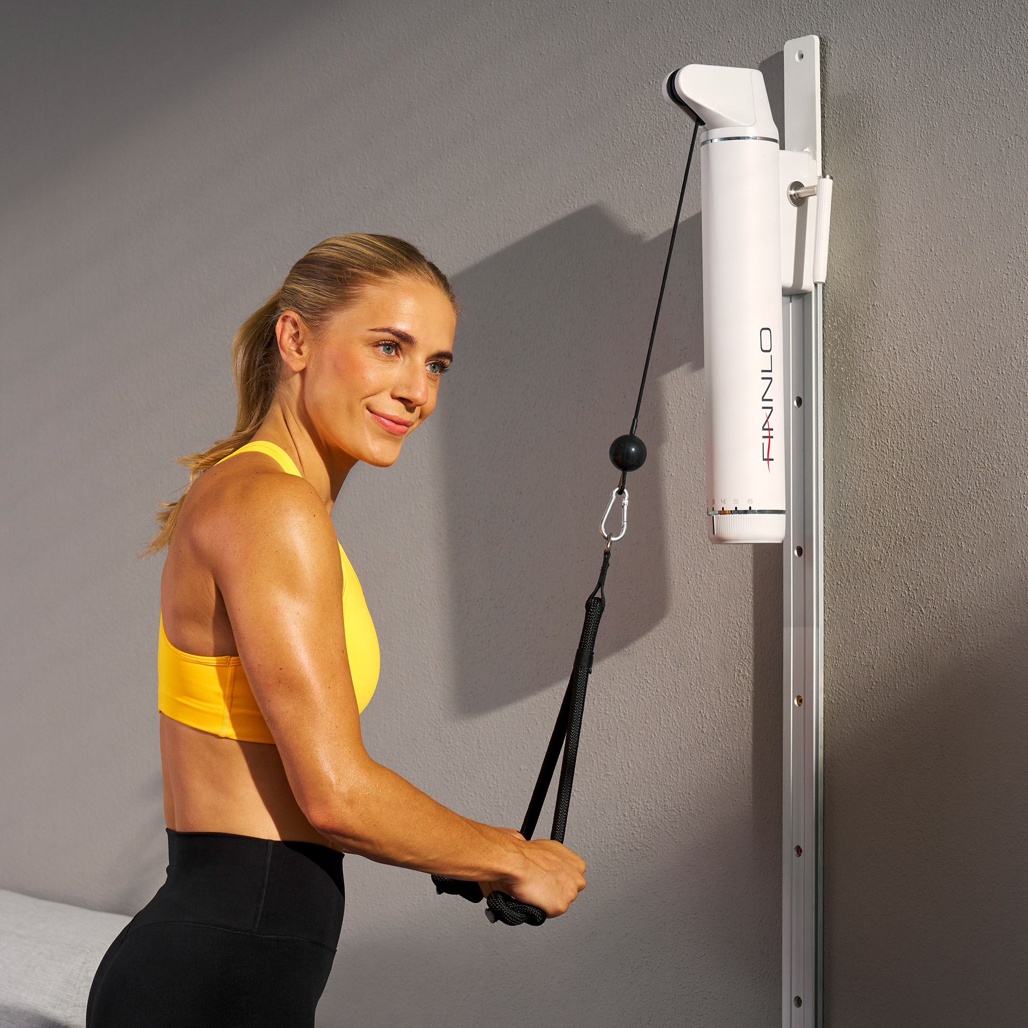 Frau in gelbem Sport-BH benutzt ein Fitnessgerät. Die Maschine ist weiß und an der Wand befestigt. Graue Wand im Hintergrund.