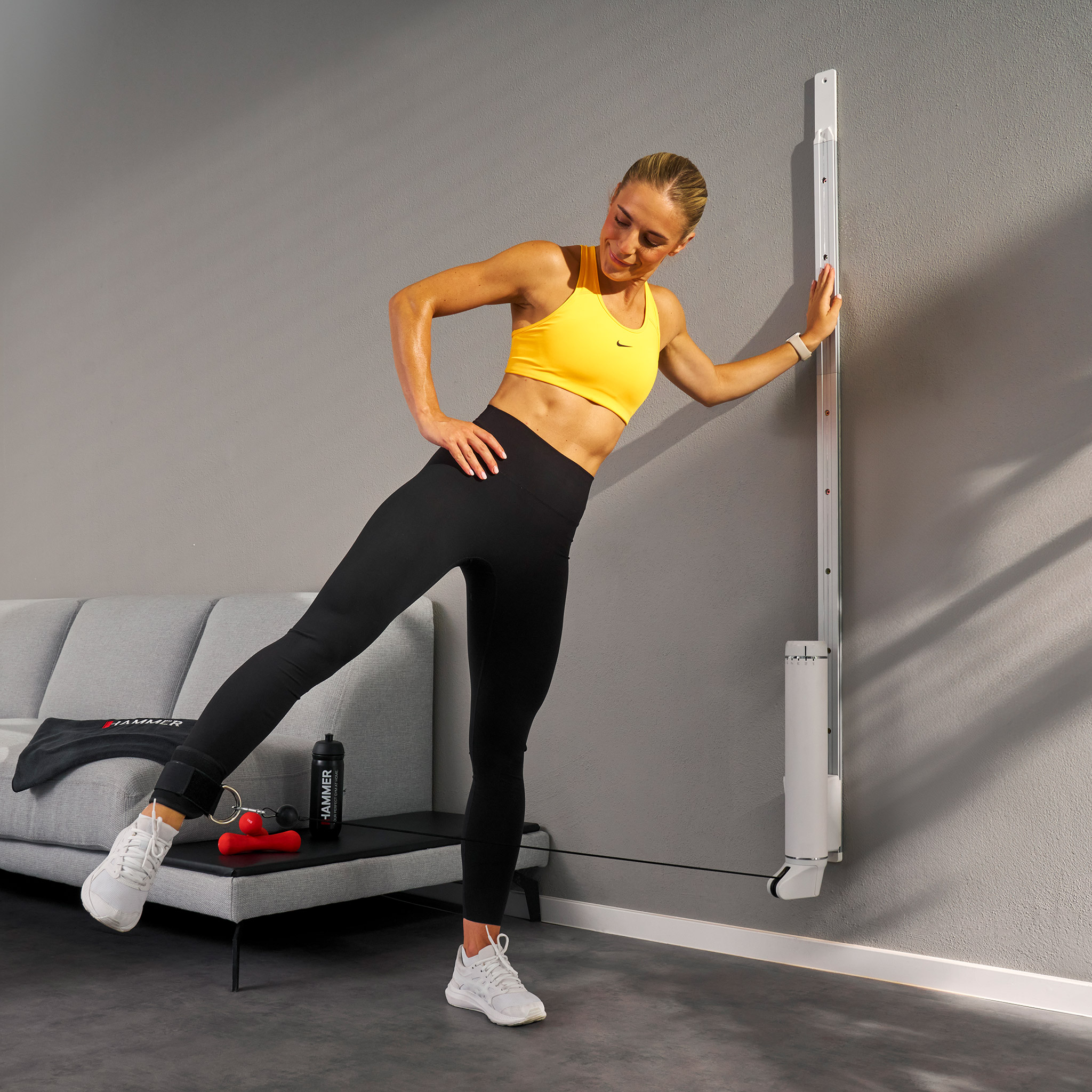 Frau in gelbem Oberteil und schwarzer Leggings macht Beinübung an der Wand. Widerstandsband an ihrem Knöchel befestigt.