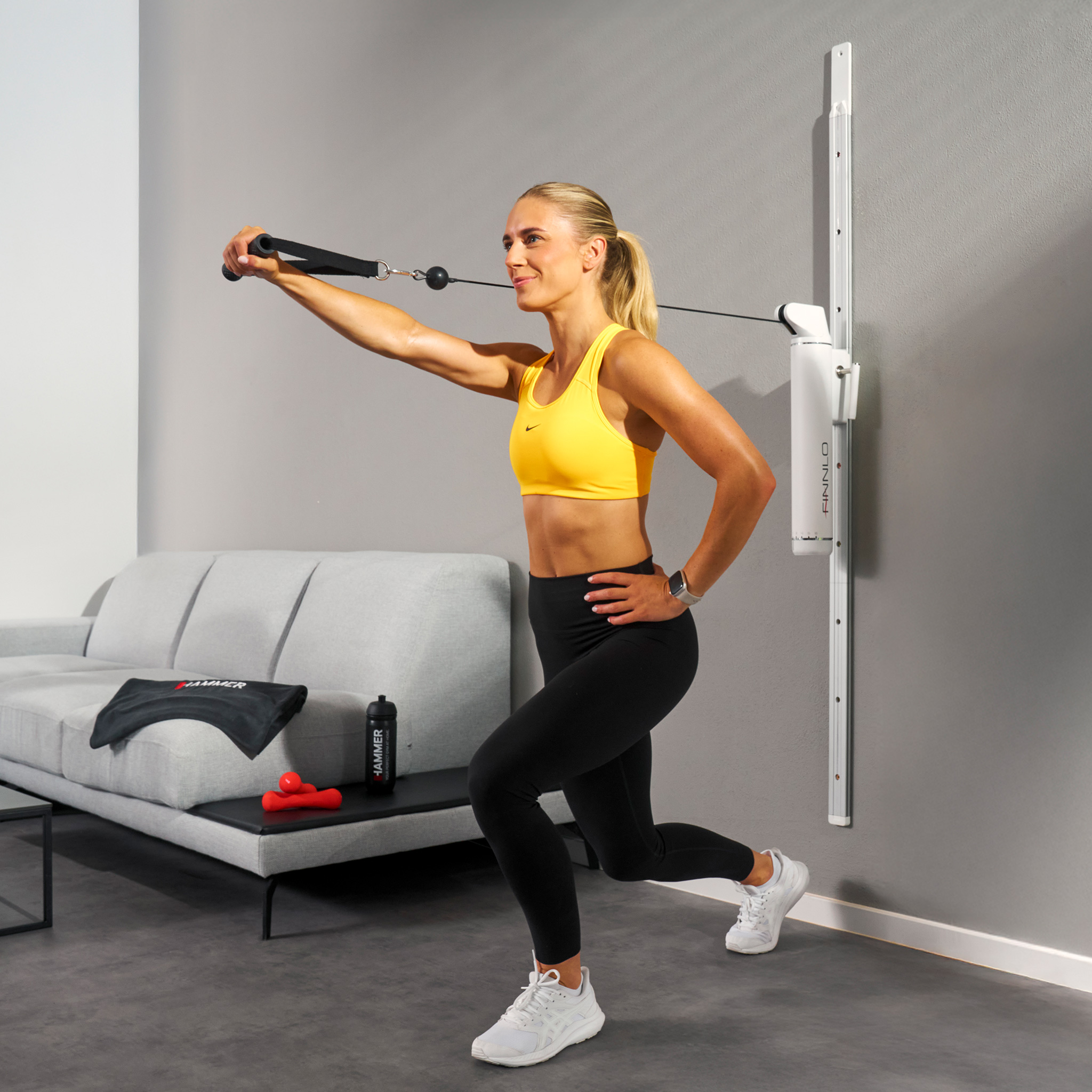Frau in gelbem Sport-BH macht Ausfallschritte mit einem wandmontierten Trainingsgerät. Graue Wand und Couch im Hintergrund.