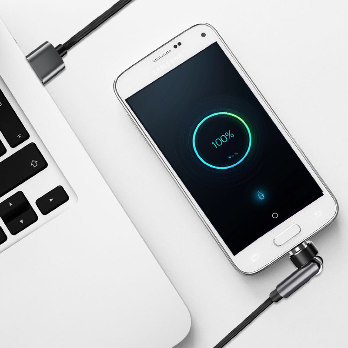 Biały telefon Samsung ładujący się za pomocą kabla podłączonego do laptopa. Ekran telefonu pokazuje 100% naładowania. Tło jest białe.
