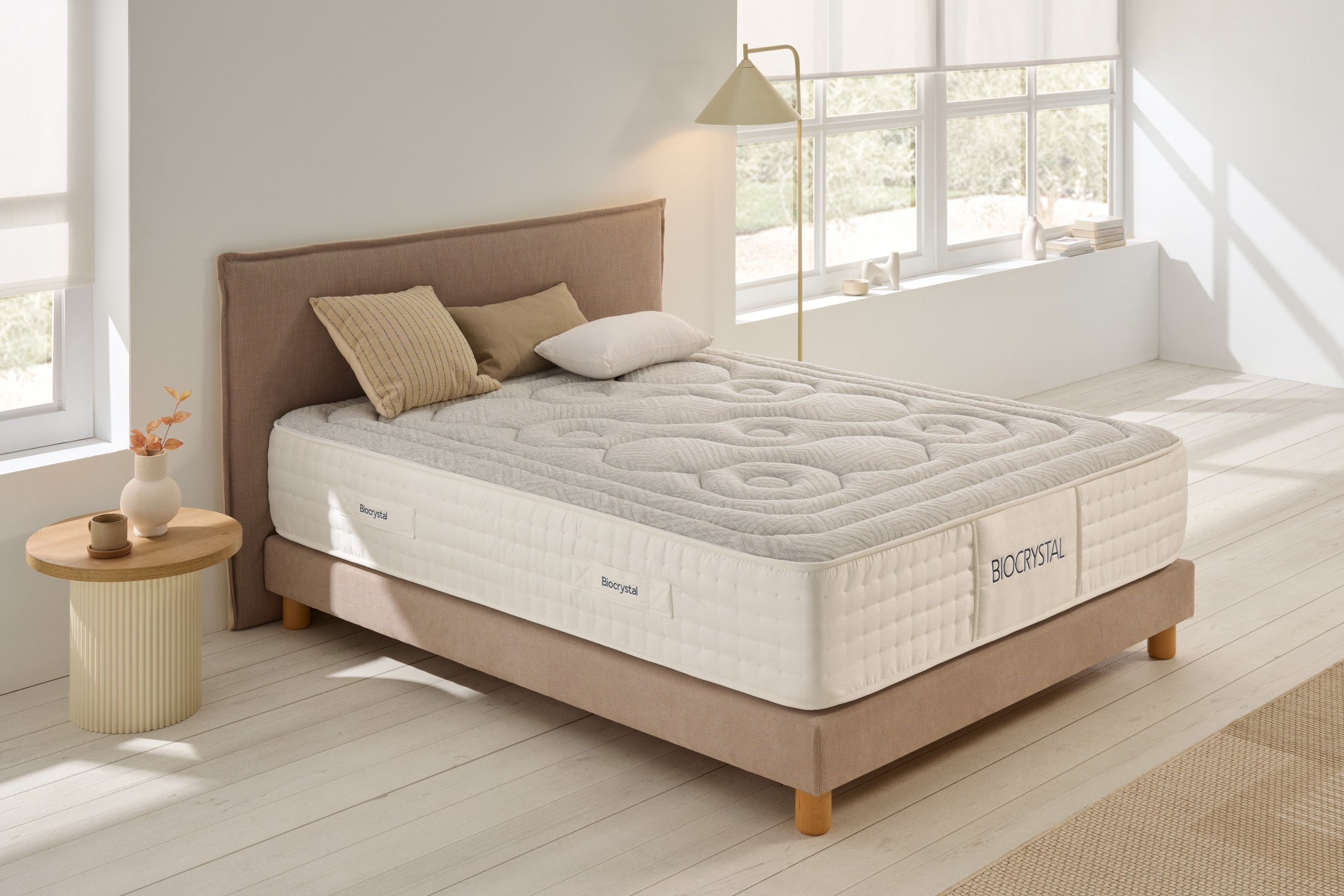 Una cama con almohadas, colchón y cabecero beige. Una mesita con un jarrón y una lámpara están al lado.