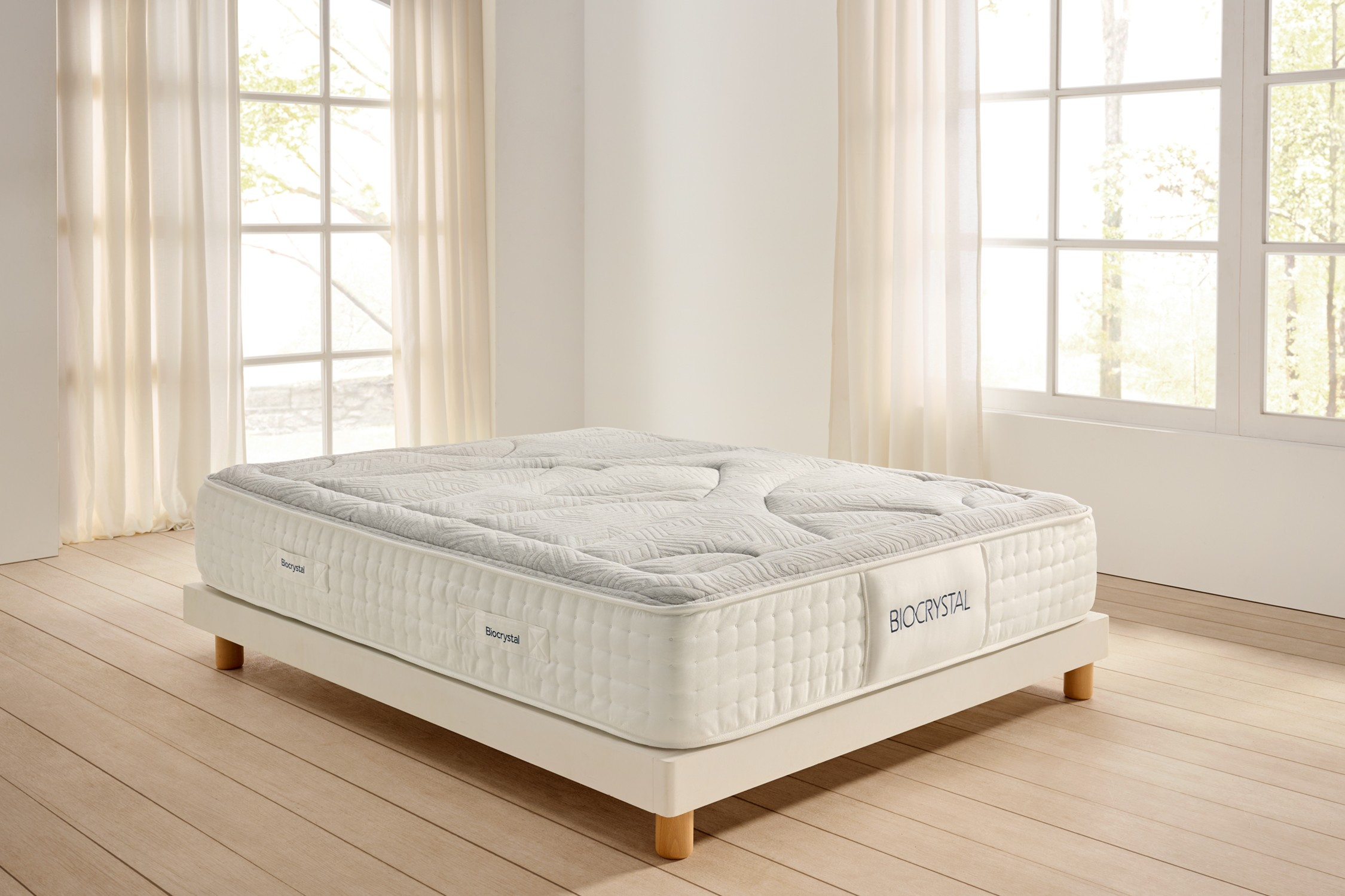 Un colchón blanco con la parte superior gris se encuentra en un marco de cama blanco. La habitación tiene pisos de madera clara y ventanas con cortinas.