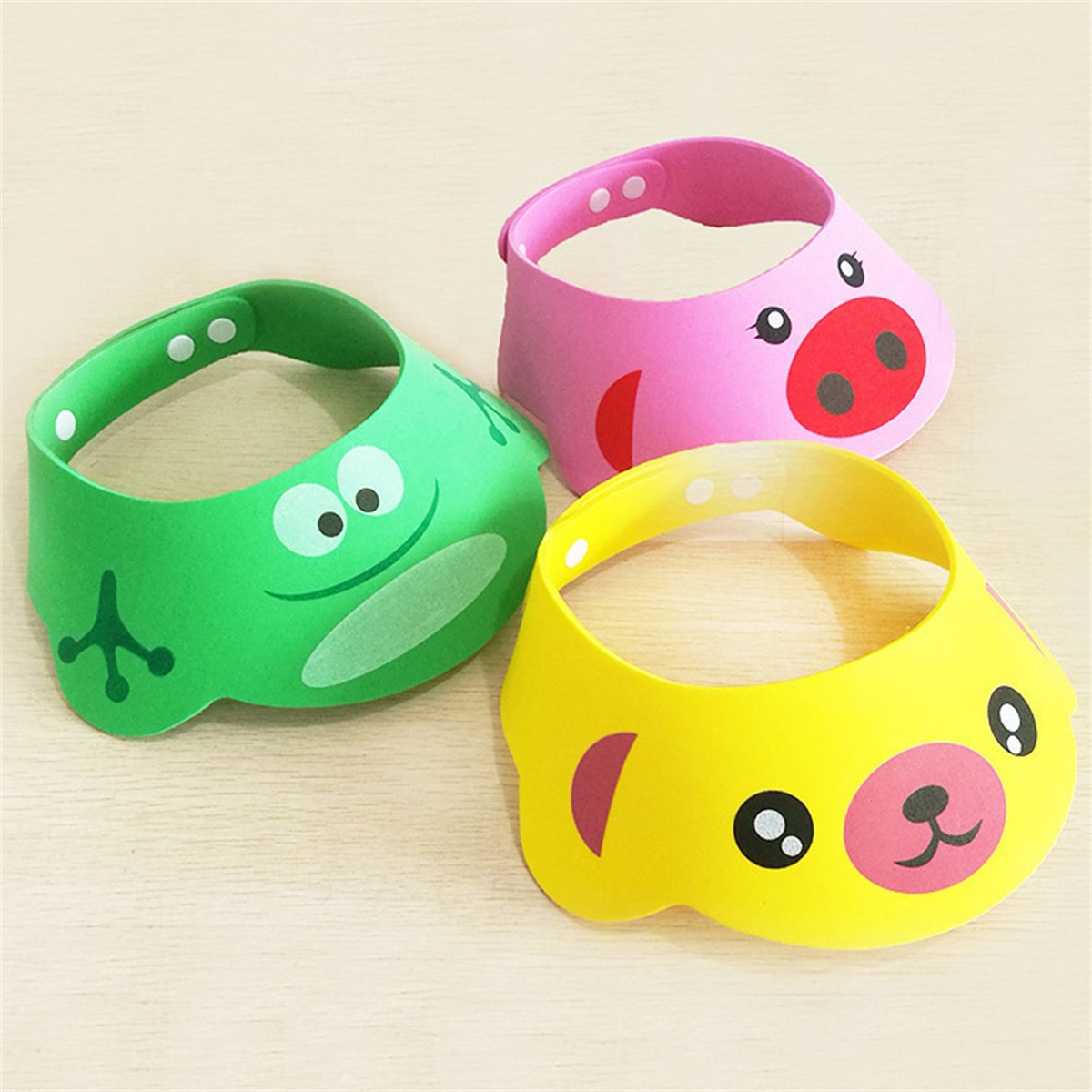 Drei bunte Badewannenvisiere: grüner Frosch, rosa Schwein und gelber Bär, angeordnet auf einer hellen Holzmaserung.