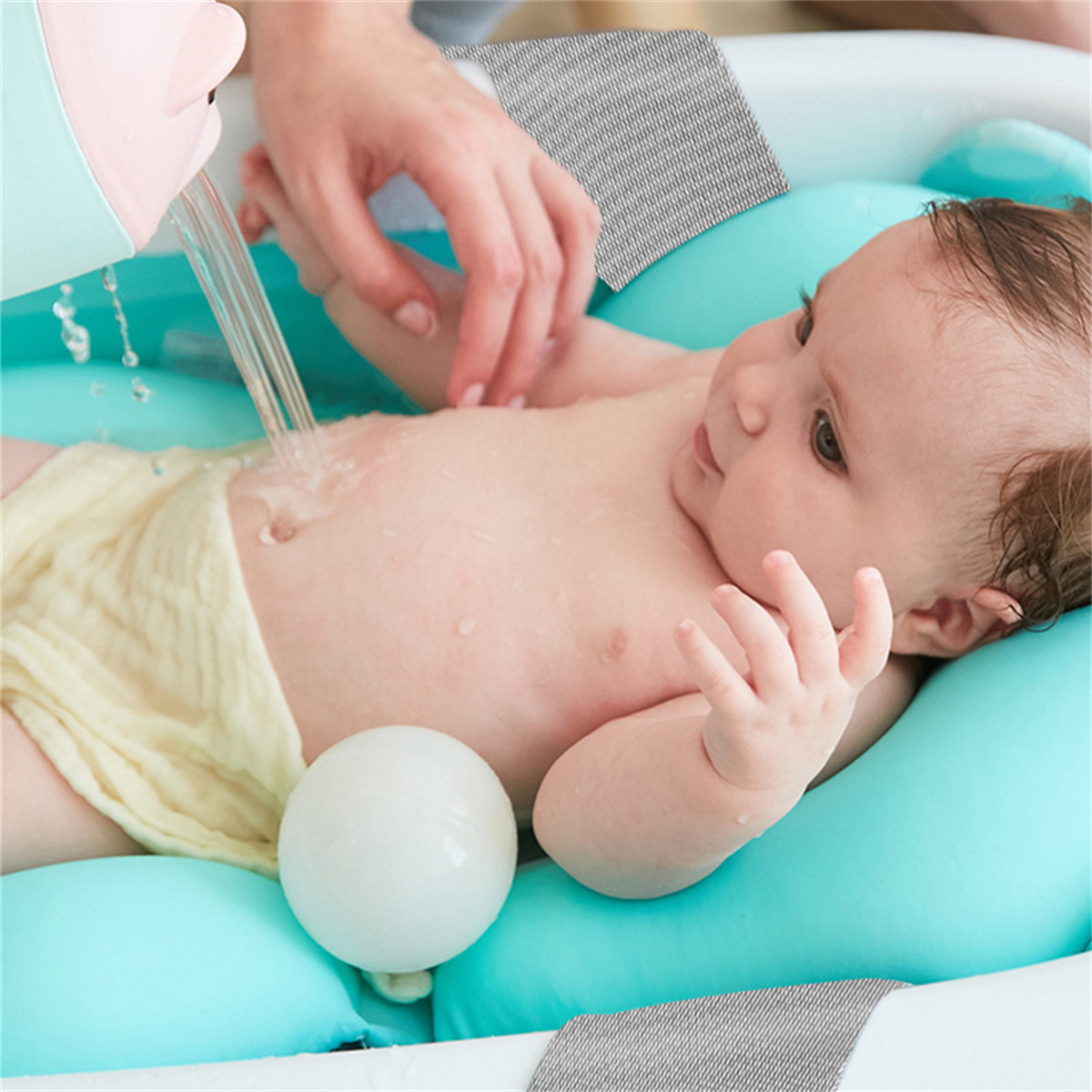 Ein Baby wird gebadet. Wasser fließt aus einem rosa Ausguss. Das Baby liegt auf einem blauen Kissen. Eine weiße Kugel ist sichtbar.