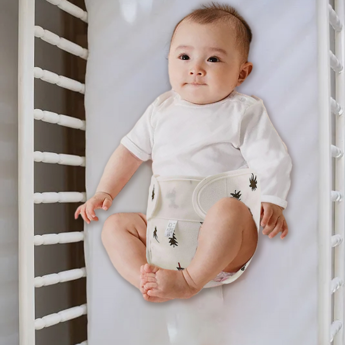 Ein Baby in einem weißen Strampler und einer beigen Windel mit schwarzen Baumdrucken, das in einem Kinderbett liegt.