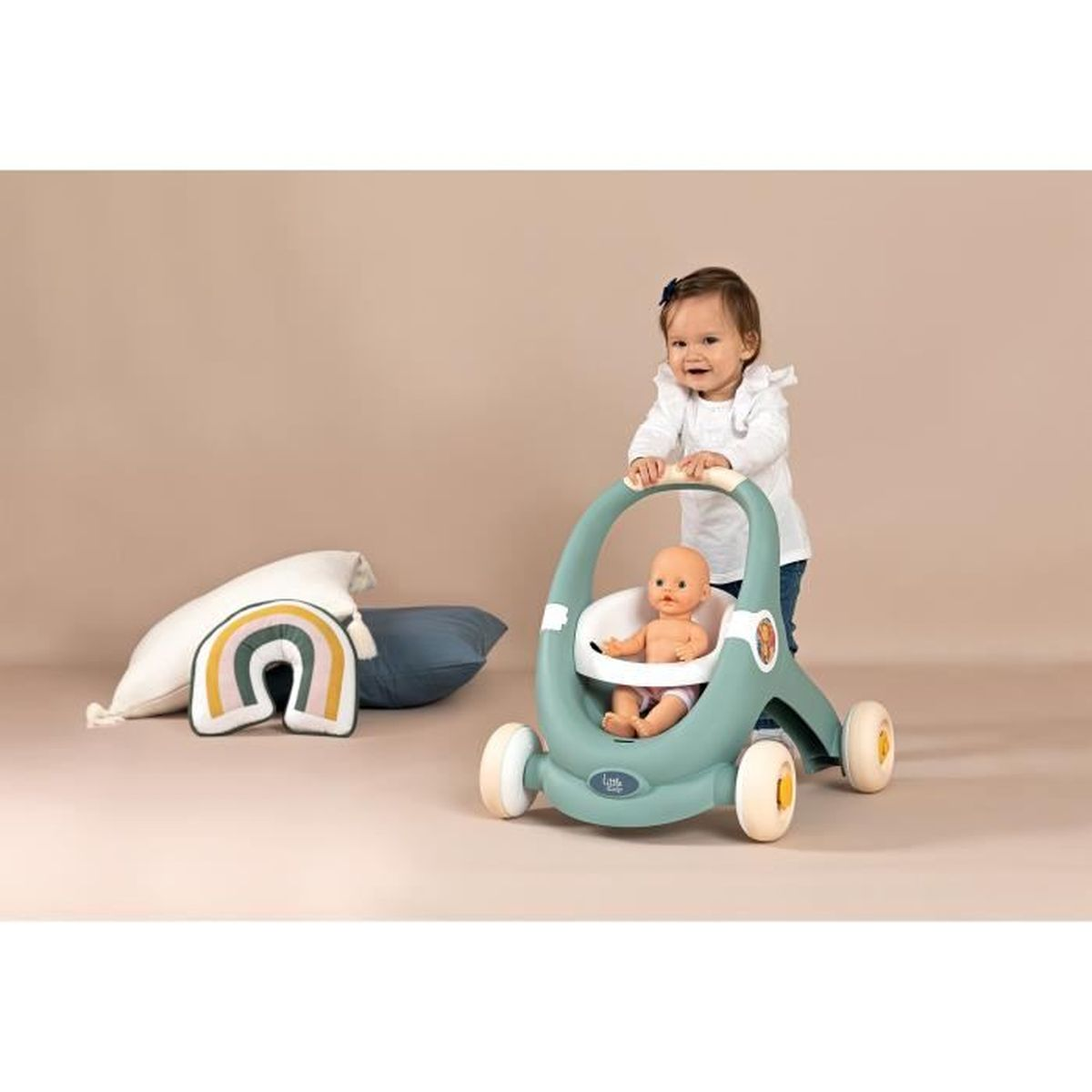 Ein Baby schiebt einen tealfarbenen Lauflernwagen mit einer Puppe darin. Kissen und ein regenbogenförmiges Kissen liegen auf dem Boden. Der Hintergrund ist schlicht.