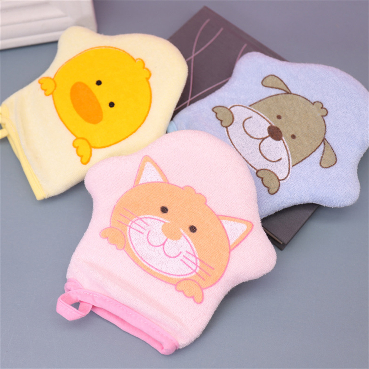 Drei farbenfrohe Waschlappen mit Tiergesichtern: Ente, Hund und Katze. Rosa, gelb und blau. Auf grauer Oberfläche mit einem Buch.