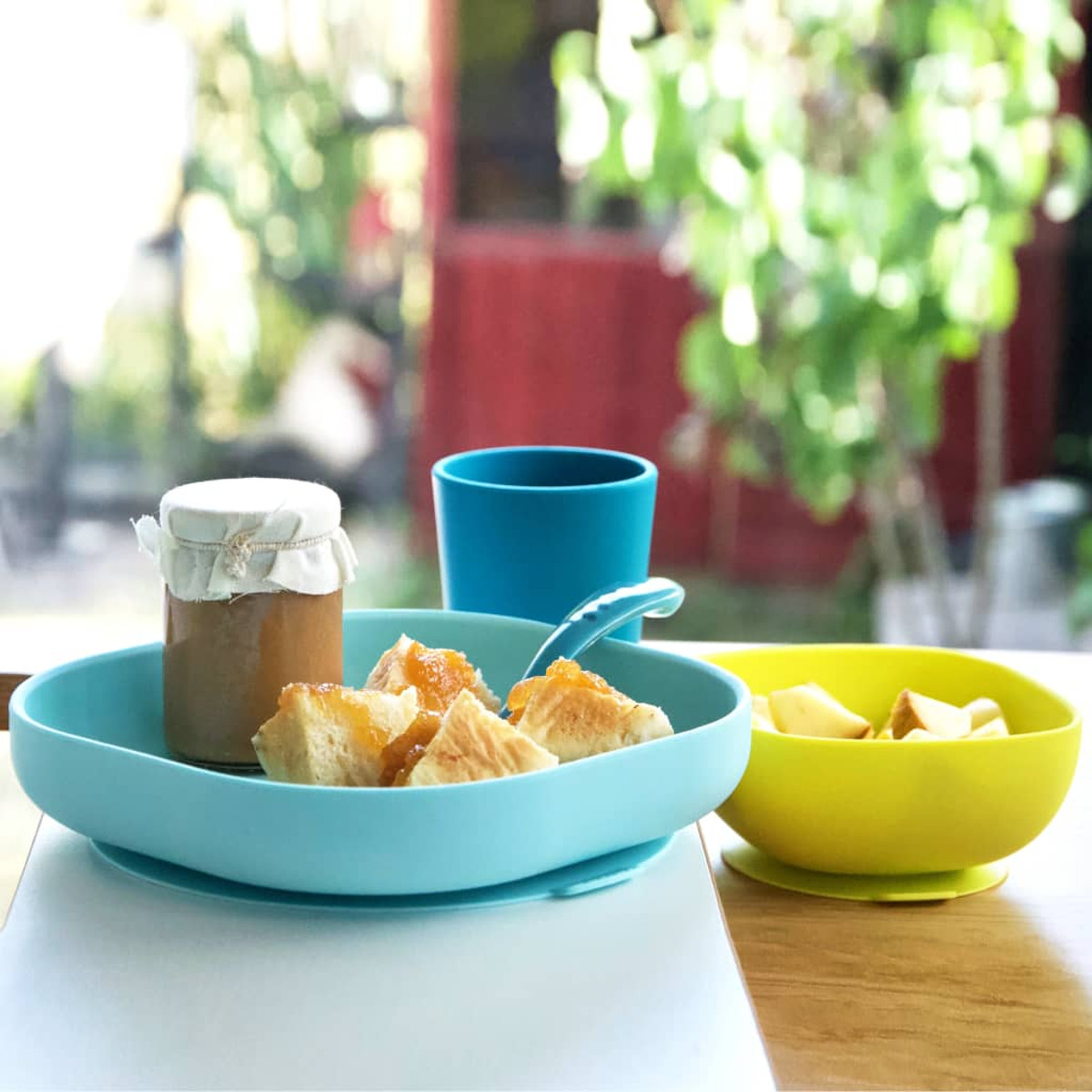 Frühstück mit Toast, Marmelade und Obst in blauen und gelben Schalen, einem blauen Becher und einem Glas. Im Hintergrund ist ein Fensterblick auf Grün.