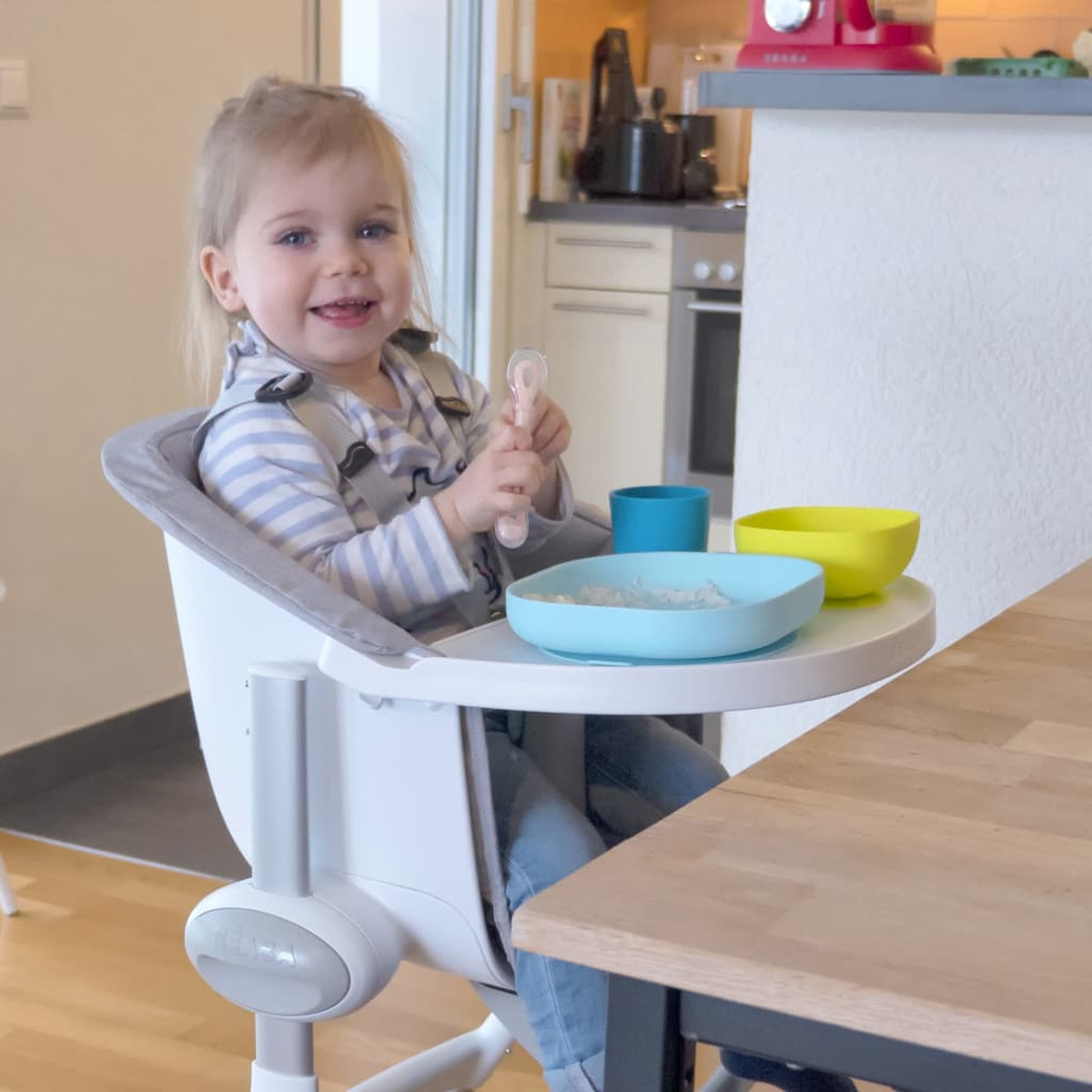 Ein lächelndes Kleinkind in einem Hochstuhl hält einen Löffel und schaut in die Kamera. Schalen mit Essen stehen auf dem Tablett. Küche im Hintergrund.