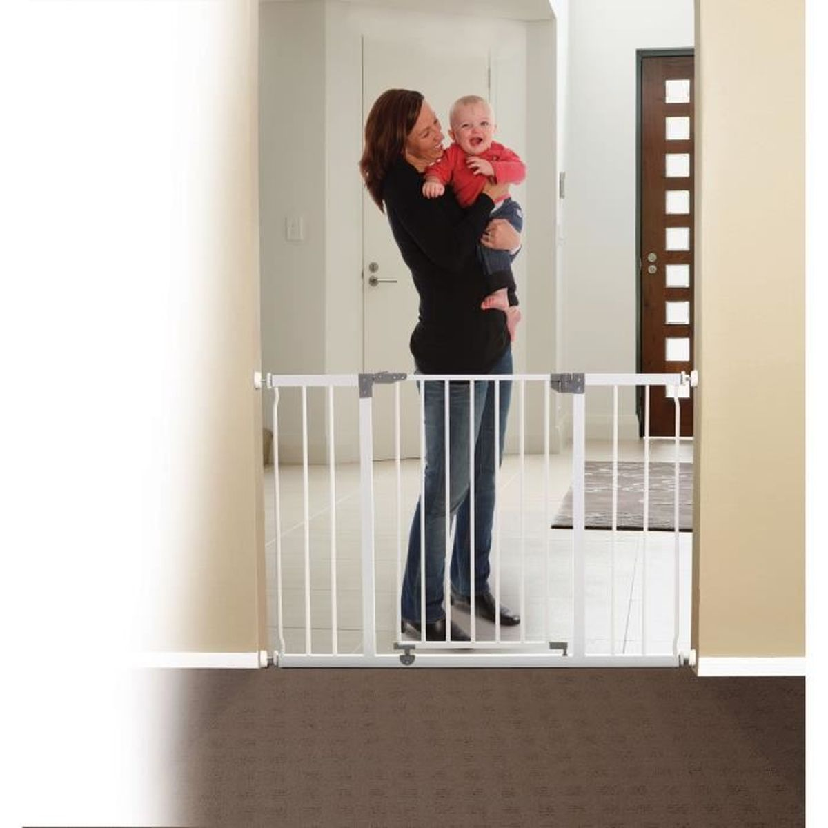 Eine Frau hält ein Baby vor einem weißen Schutzgitter. Das Baby trägt ein rotes Hemd. Der Hintergrund zeigt eine Tür und einen Flur.