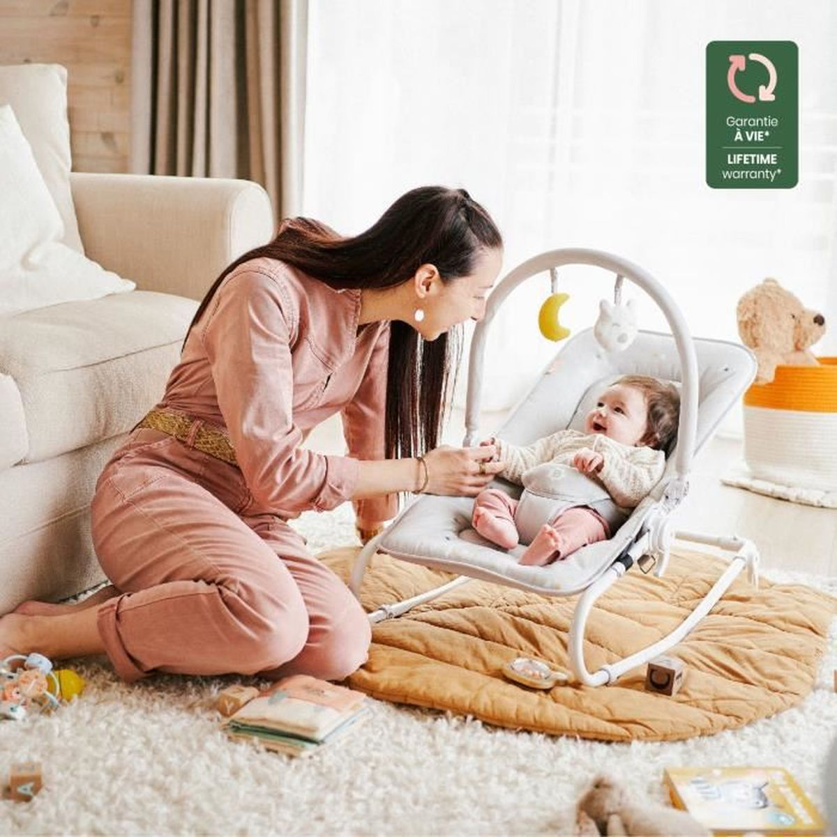Eine Frau interagiert mit einem Baby in einer Wippe. Das Baby lächelt. Ein Teddybär und ein Korb sind im Hintergrund.