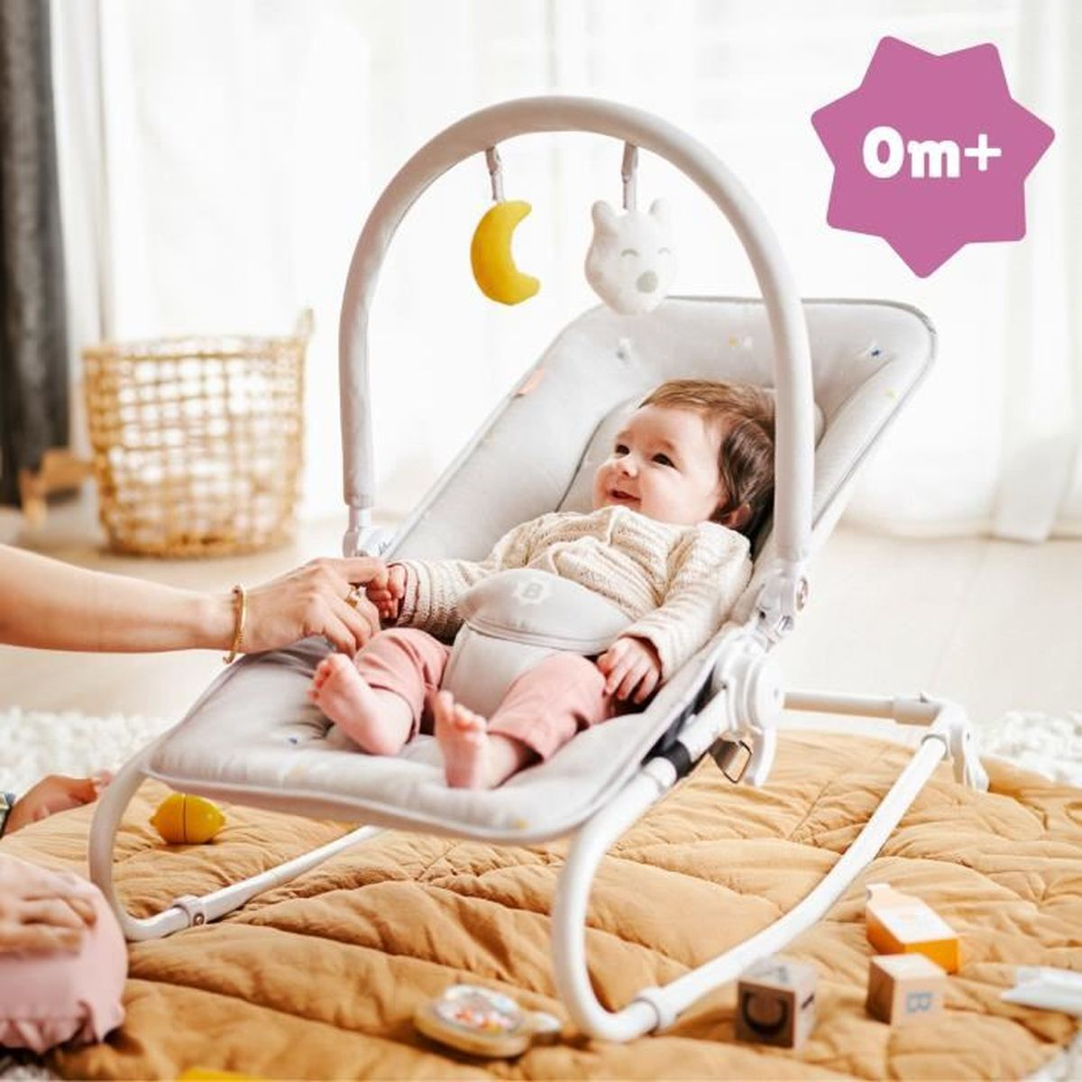 Ein Baby in einer grauen Wippe mit Mond- und Bärspielzeug. Eine Hand hält die Hand des Babys. 0m+ Zeichen sichtbar.