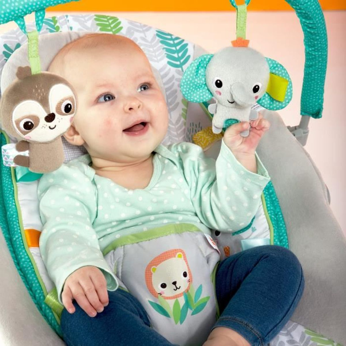 Ein Baby in einer Wippe lächelt und blickt nach oben. Spielzeug: Faultier, Elefant. Der Bouncer hat grüne Akzente und ein Löwenmuster.