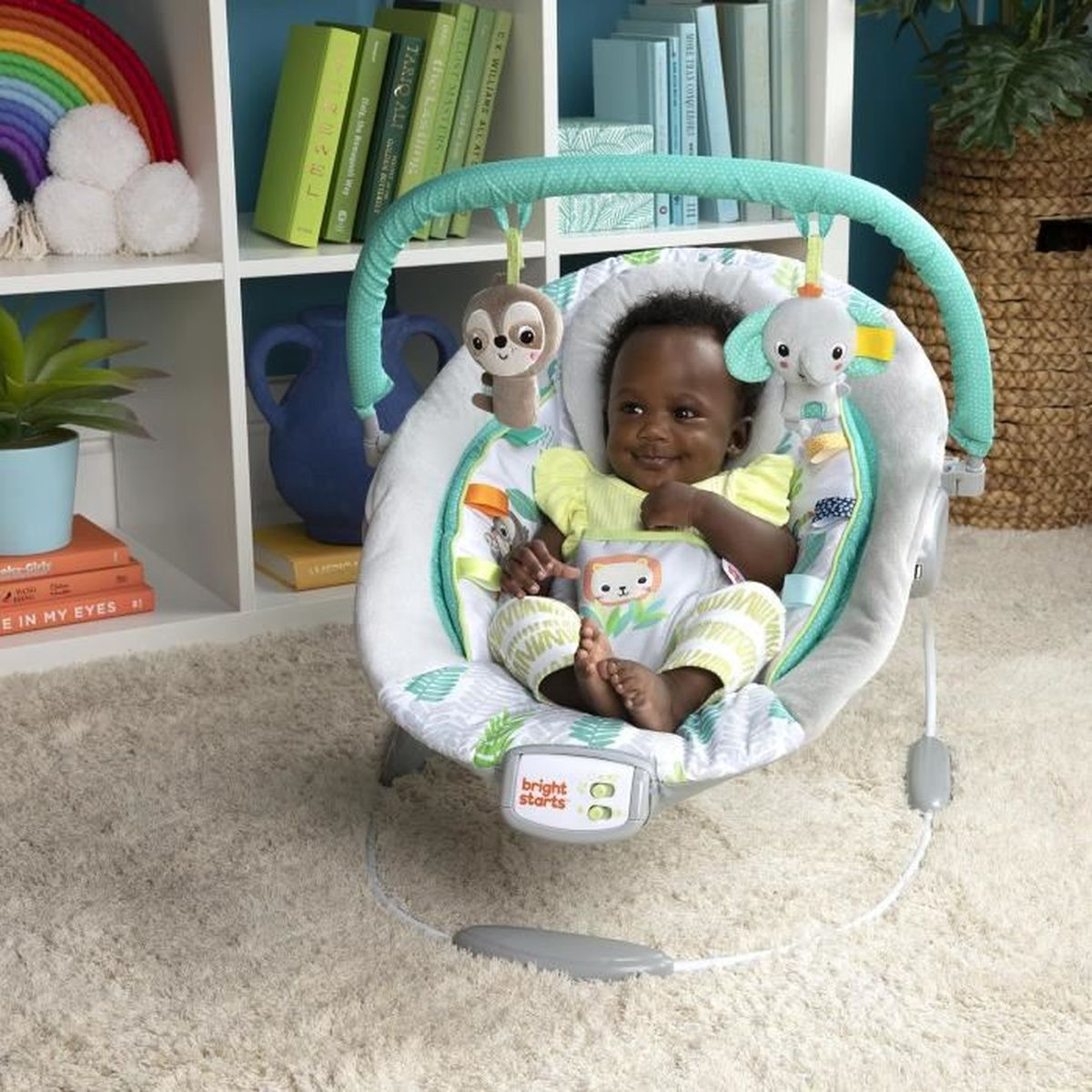 Ein Baby sitzt in einer Wippe mit Spielzeug. Regale halten Bücher und Dekorationen. Das Baby lächelt.