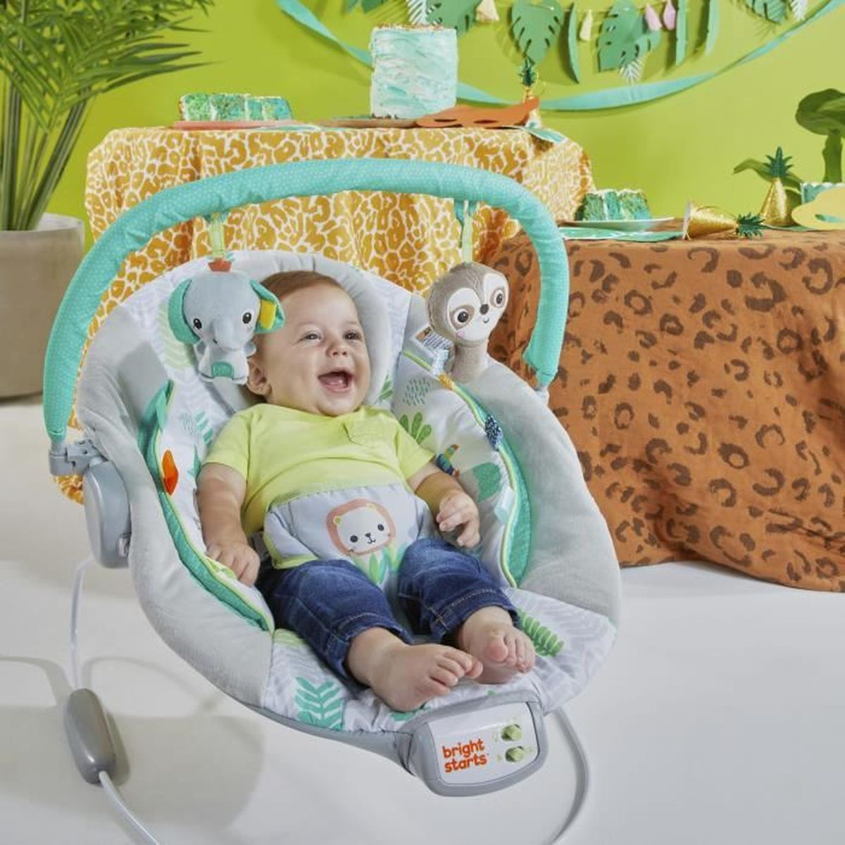 Ein Baby lacht in einer Wippe mit Spielzeug. Die Szene hat Dschungeldekorationen, eine Tischdecke mit Giraffenmuster und eine Tischdecke mit Leopardenmuster.