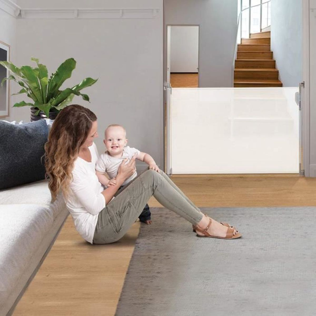 Eine Frau und ein Baby sitzen in der Nähe eines Sicherheitsgitters. Links sind eine Pflanze und eine Couch. Im Hintergrund ist eine Treppe. Das Baby lächelt.