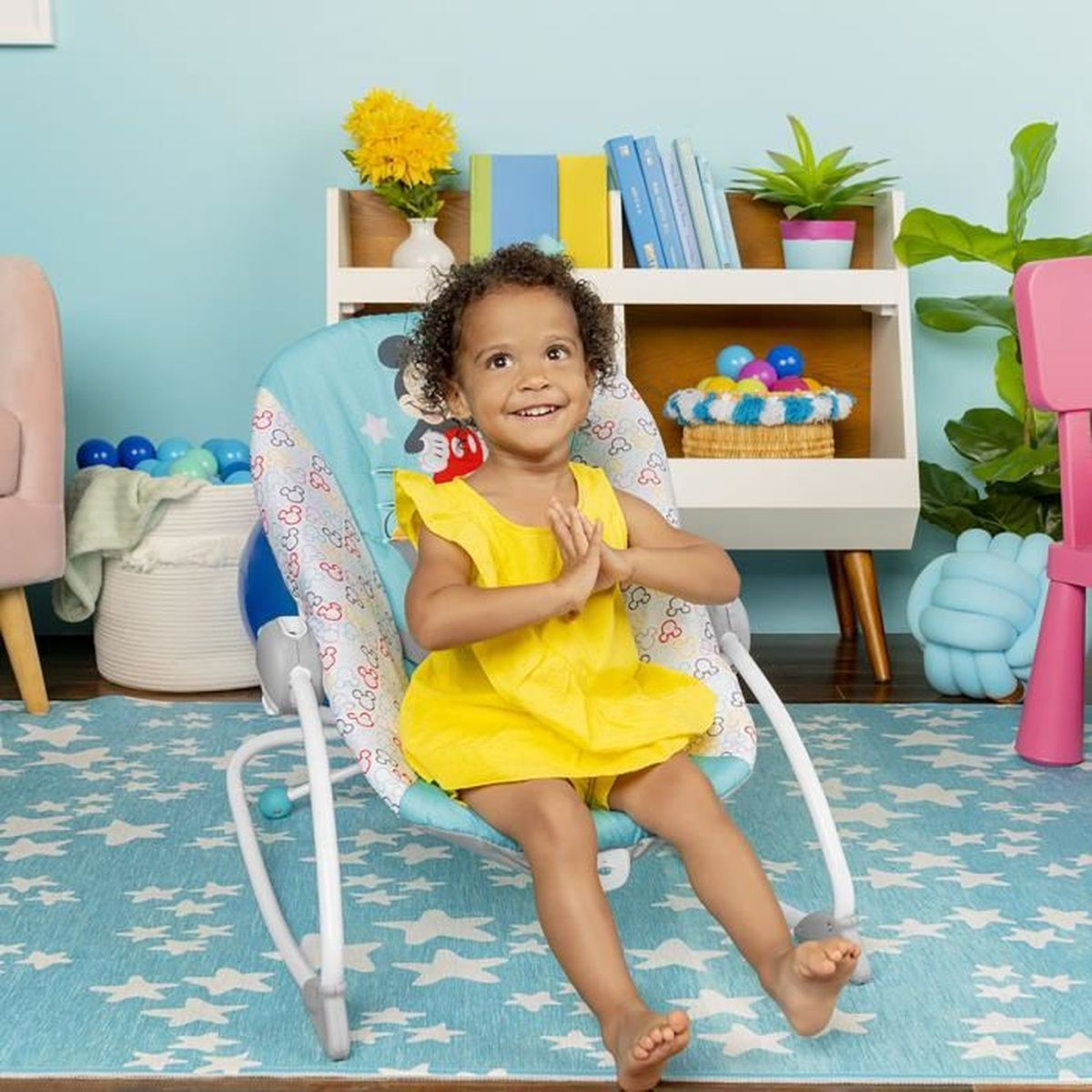 Ein lächelndes Kind in einem gelben Kleid sitzt in einer Wippe. Der Hintergrund hat eine blaue Wand, ein Bücherregal und einen Teppich mit Sternenmuster.