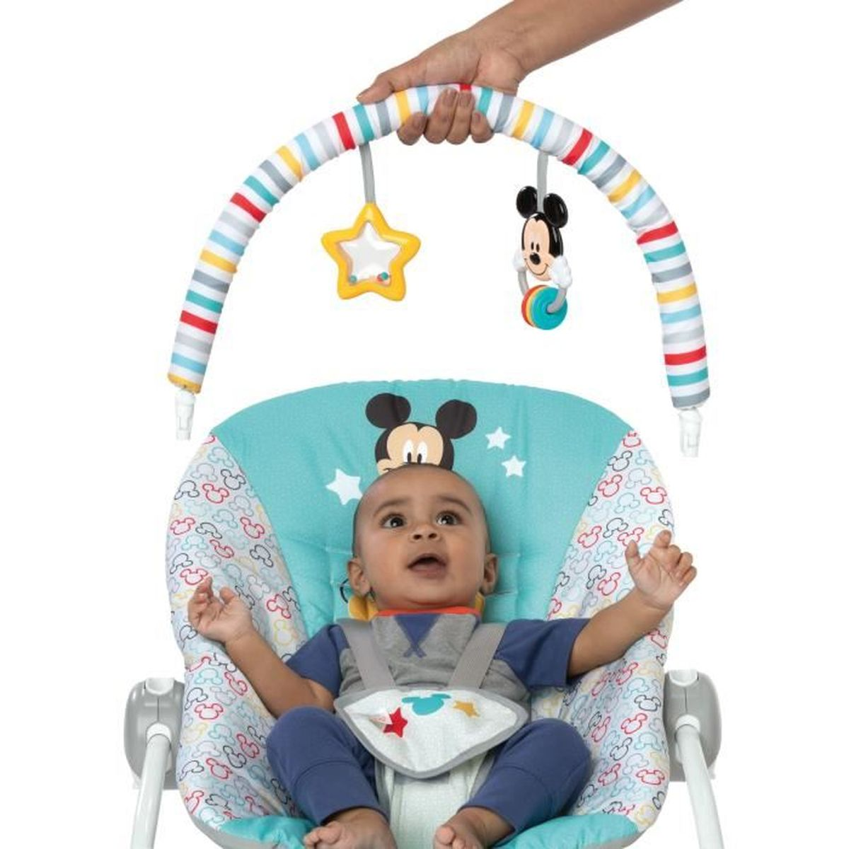 Ein Baby liegt in einer Wippe mit Mickey Mouse-Motiv. Über dem Baby hängen Spielzeuge. Eine Hand hält den Spielzeugbogen.