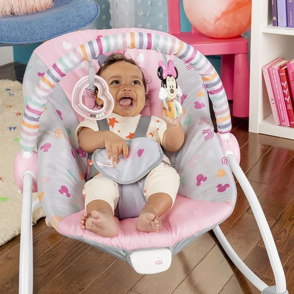Ein Baby sitzt in einer rosa-grauen Wippe, lächelt und hält ein Minnie-Maus-Spielzeug. Im Hintergrund ein Regal mit Büchern.