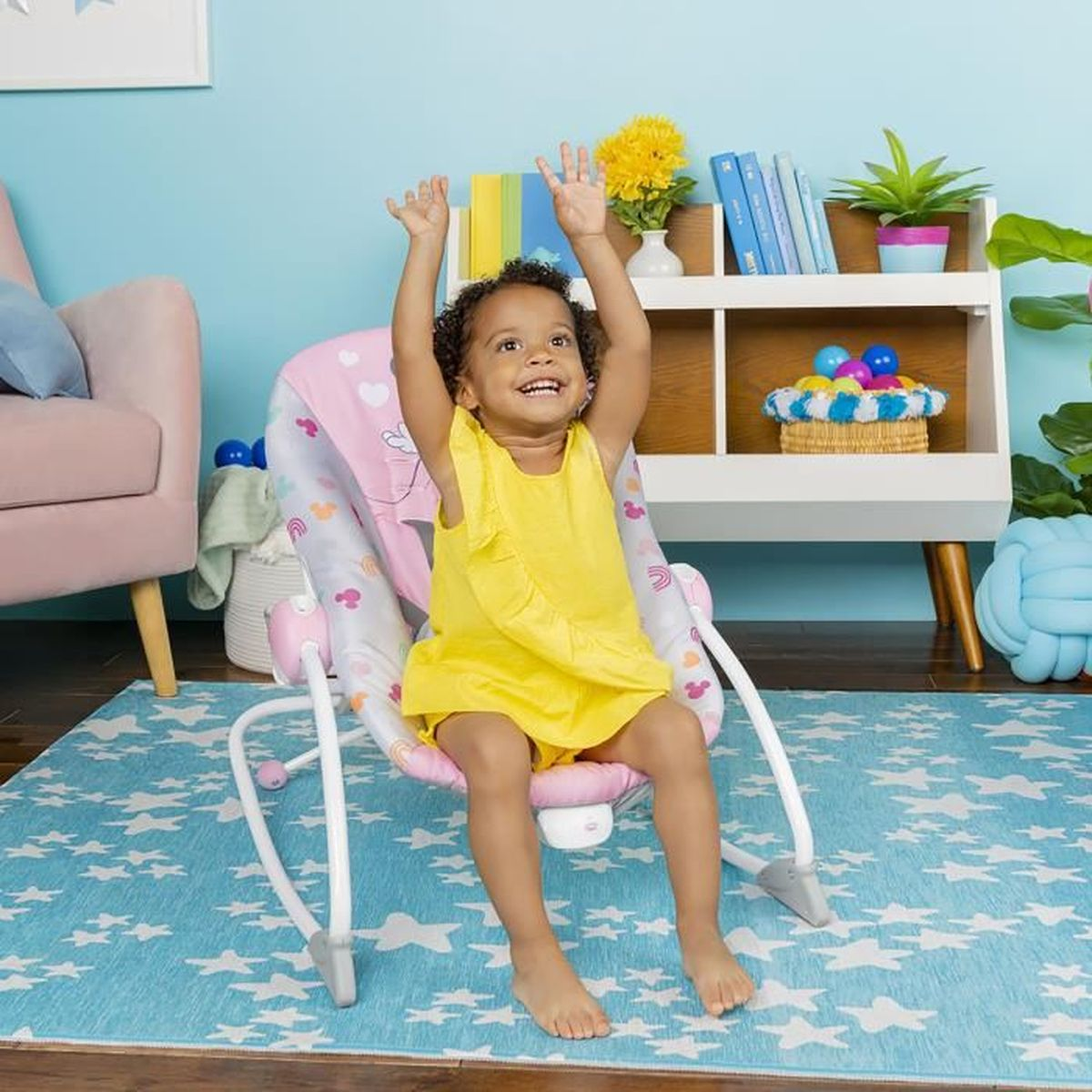 Ein lächelndes Kleinkind in einem gelben Kleid sitzt in einer rosa-weißen Babywippe mit erhobenen Armen. Der Hintergrund umfasst Möbel und einen blauen Teppich.