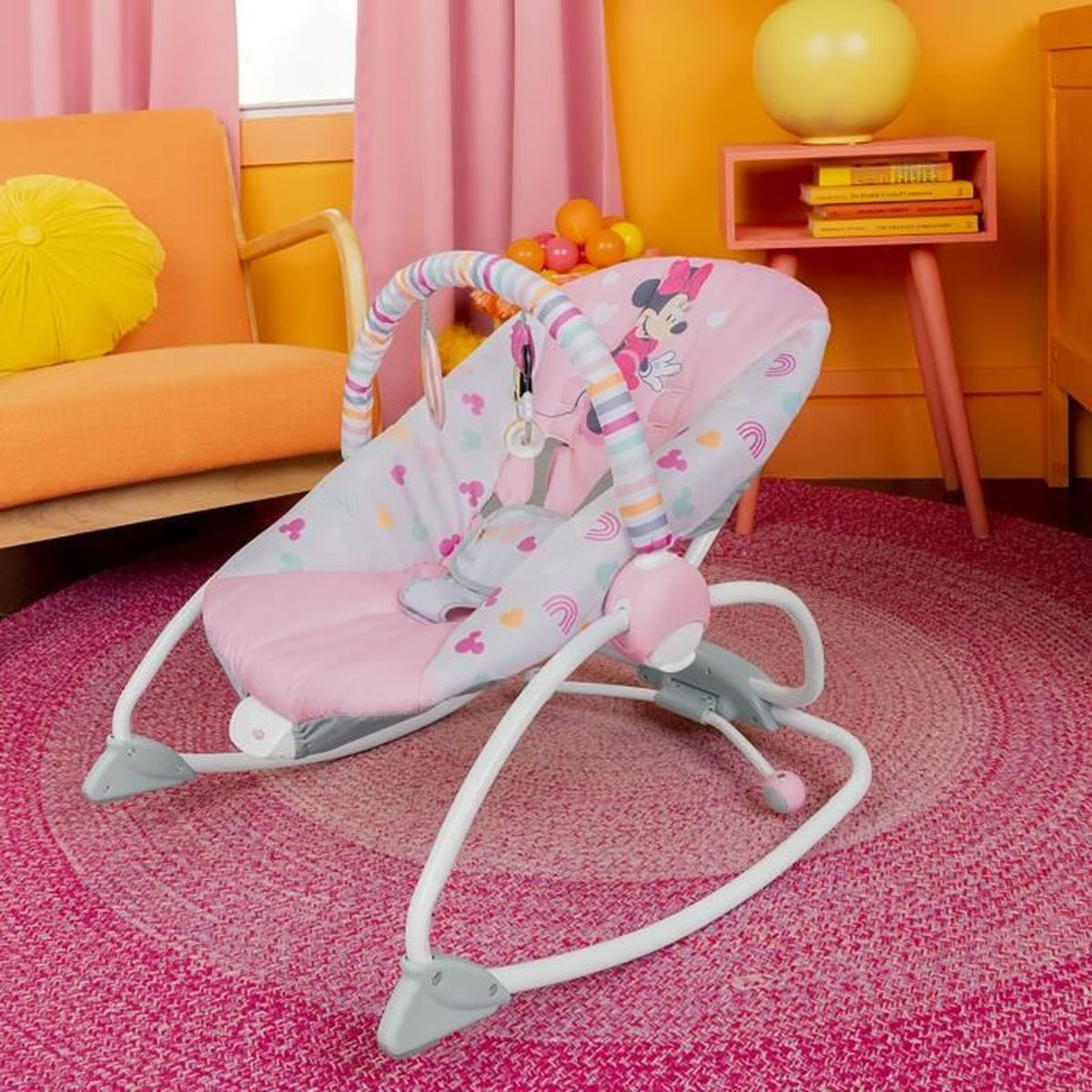 Eine Babywippe in einem rosa Zimmer. Sie hat ein Minnie-Maus-Design. Im Hintergrund sind ein rosa Teppich und ein oranger Stuhl.