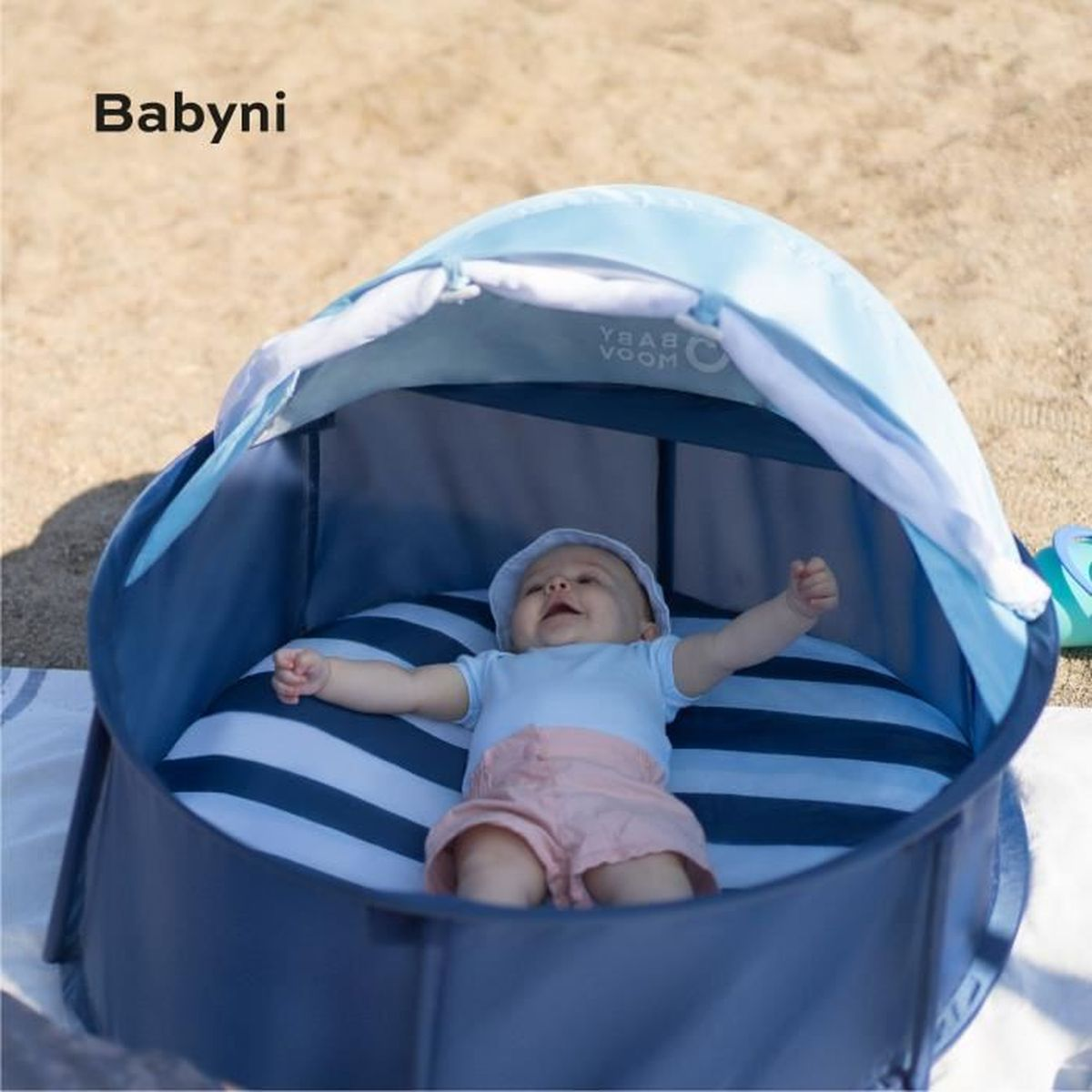 Ein Baby liegt in einem blau-weiß gestreiften Spielzelt am Strand. Das Baby trägt einen hellblauen Anzug und rosa Shorts.