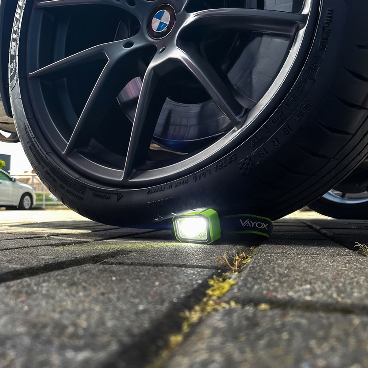 Zielono-czarna lampa robocza Vayox znajduje się pod oponą samochodu. Opona i koło są czarne. W tle widać biały samochód.