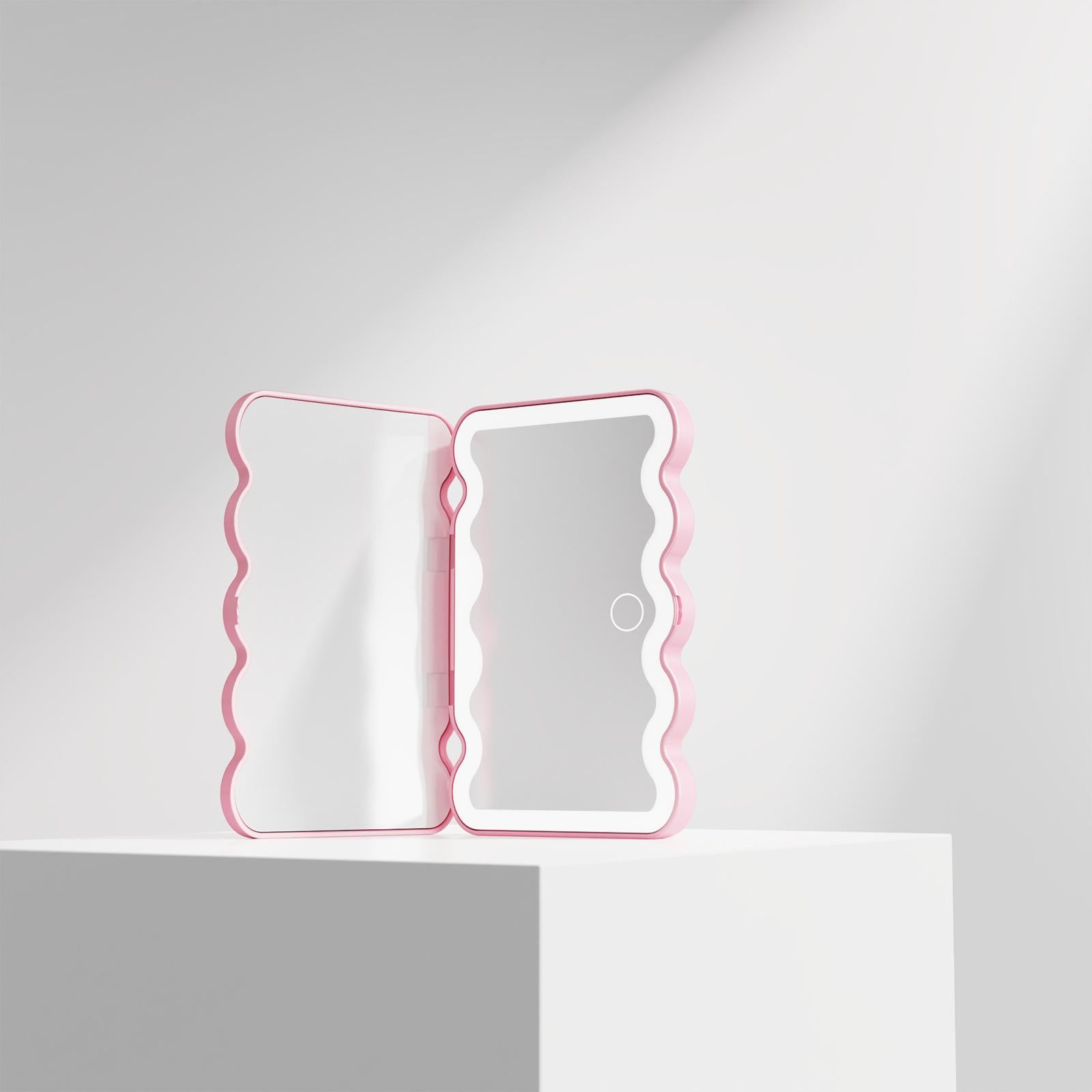 Ein rosa, wellenförmiger, offener Spiegel steht auf einer weißen Oberfläche. Der Spiegel ist geöffnet und zeigt zwei reflektierende Flächen.