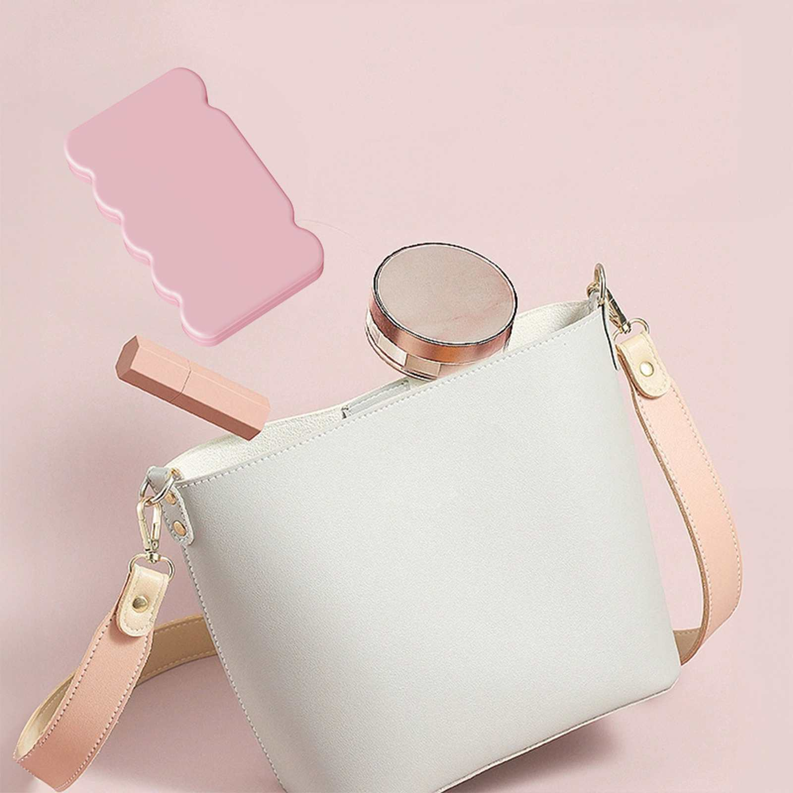 Eine weiße Handtasche mit rosa Riemen, eine rosa Gesichtswalze, Lippenstift und ein Kompaktpuder auf rosa Hintergrund.