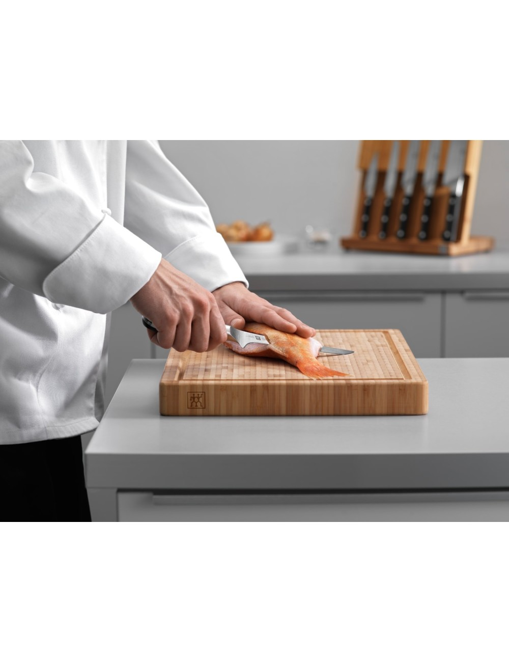 Szef kuchni kroi czerwoną rybę na drewnianej desce do krojenia. Noże w tle. Szef kuchni ma na sobie biały fartuch.