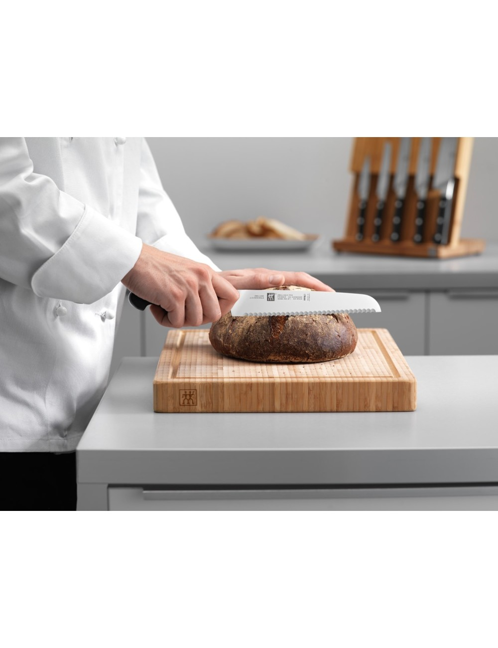 Osoba kroi bochenek chleba na drewnianej desce z ząbkowanym nożem. Kilka noży znajduje się w drewnianym bloku w tle.