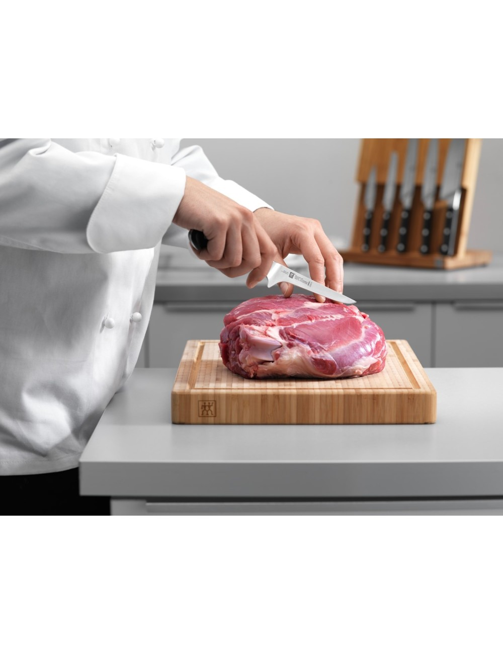 Szef kuchni kroi surowe mięso na drewnianej desce do krojenia. Noże znajdują się w drewnianym bloku w tle. Scena rozgrywa się w kuchni.