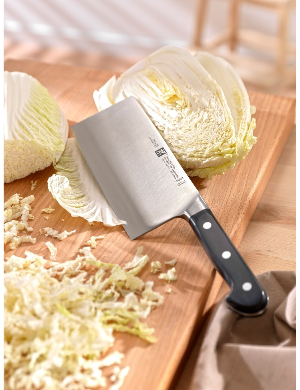 Ein Hackmesser liegt auf einem Holzbrett mit geschnittenem und ganzem Kohl. Der Hintergrund zeigt einen Holztisch und ein Tuch.