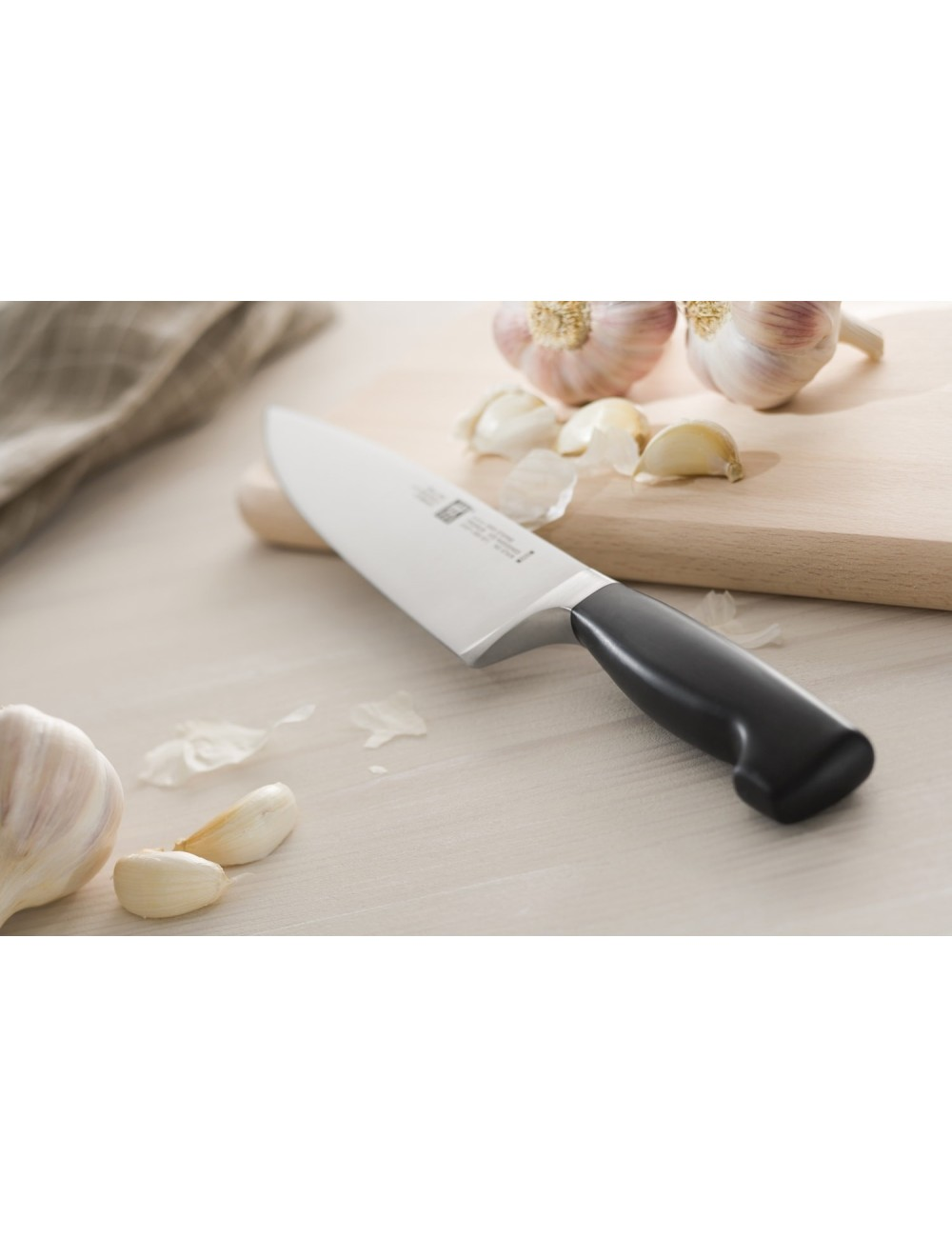 Nóż z czarnym uchwytem na jasnej drewnianej powierzchni, z ząbkami i główkami czosnku. Drewniana deska do krojenia w tle.
