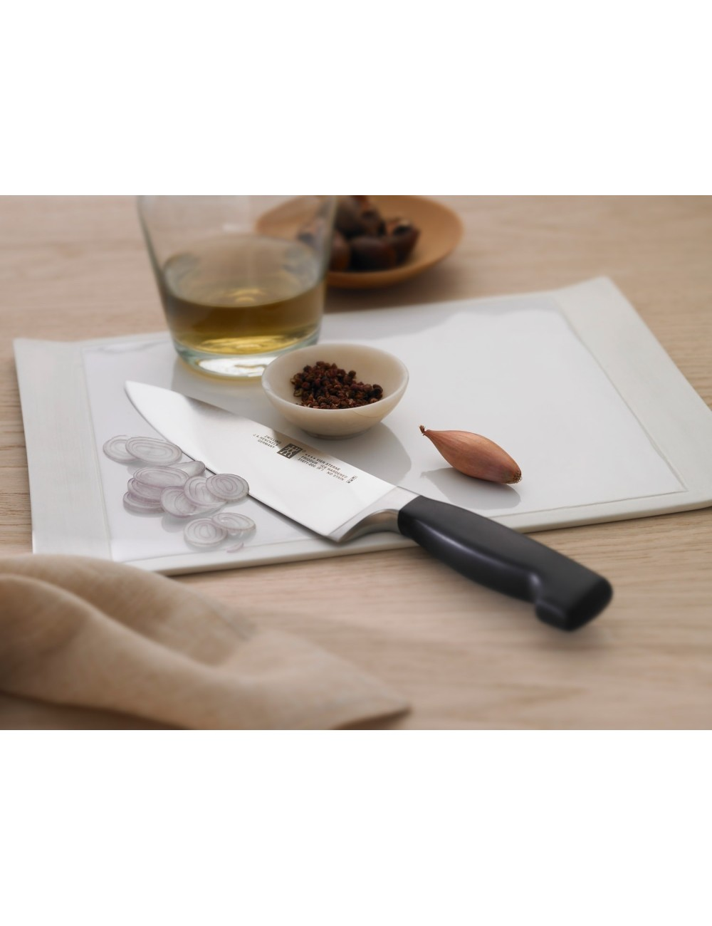 Nóż spoczywa na białej desce do krojenia z pokrojoną cebulą, szalotką i małymi miseczkami z przyprawami. W pobliżu stoi szklanka płynu.