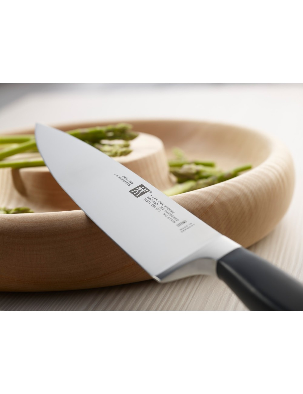 Zbliżenie noża szefa kuchni z czarną rączką, spoczywającego na drewnianej misce ze szparagami. Nóż jest oznaczony.