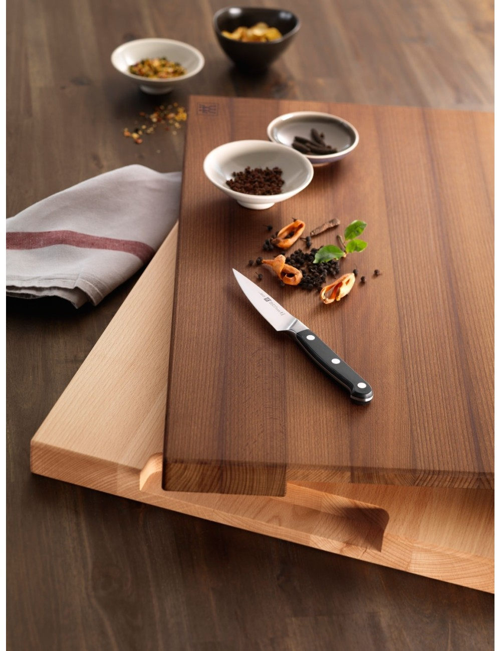 Nóż spoczywa na drewnianej desce do krojenia z przyprawami i małymi miseczkami. Ściereczka leży z boku, na jaśniejszej drewnianej desce.