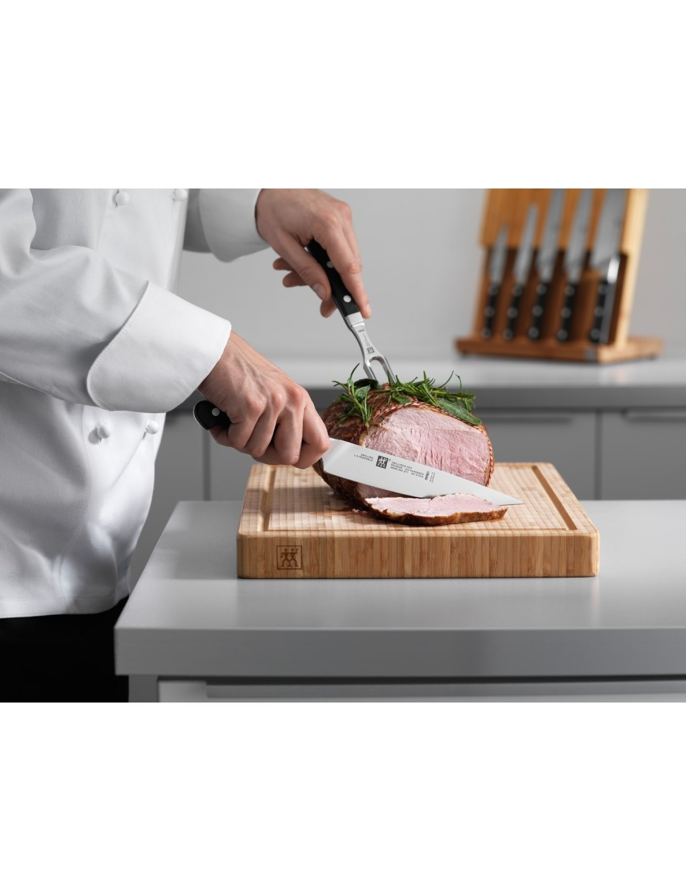 Szef kuchni kroi pieczone mięso na drewnianej desce do krojenia nożem i widelcem. Zestaw noży jest w tle.