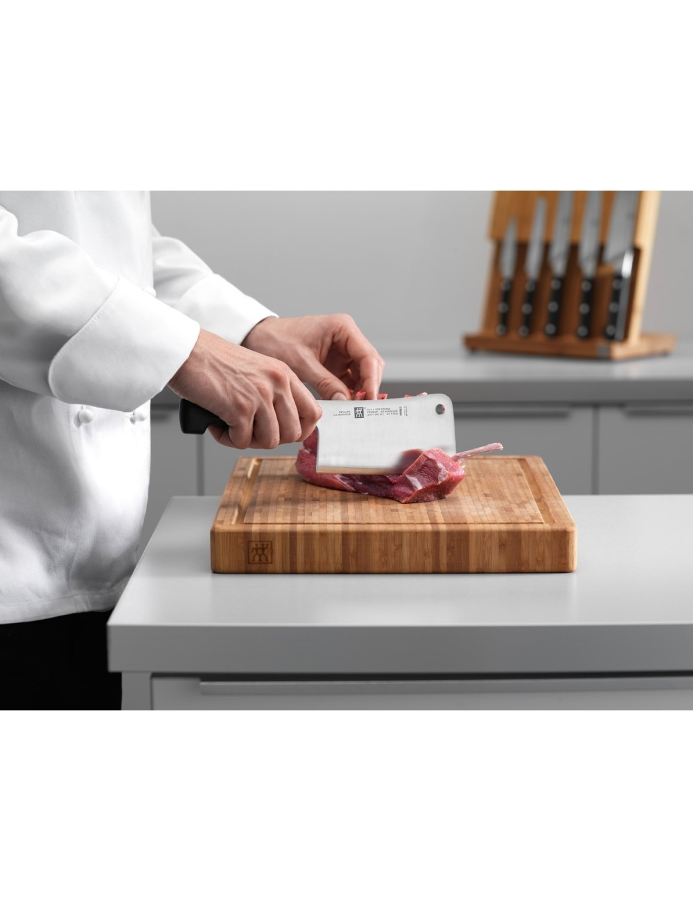 Szef kuchni kroi mięso na drewnianej desce tasakiem. Noże znajdują się w drewnianym bloku w tle. Scena rozgrywa się w kuchni.