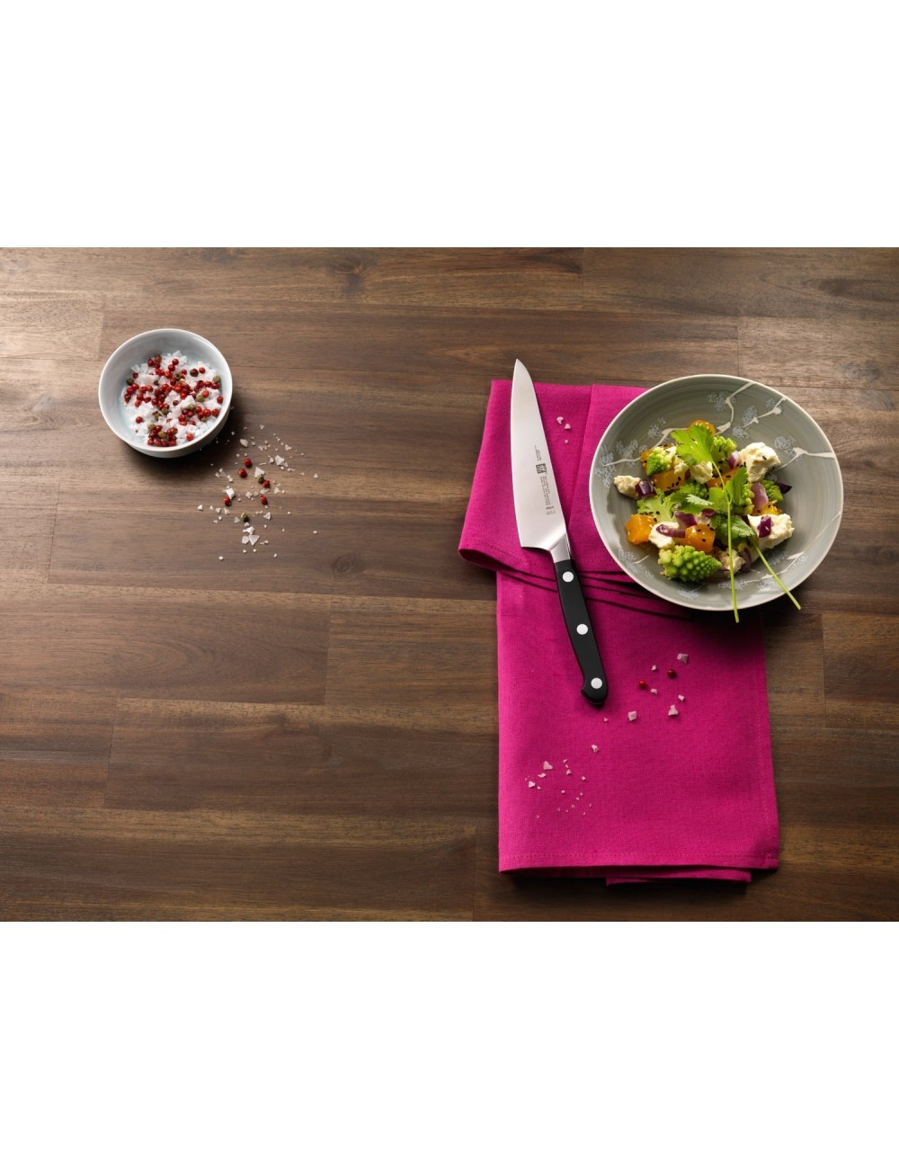 Nóż spoczywa na różowej serwetce obok miski z sałatką. Po lewej stronie mała miska z przyprawami. Wszystko na drewnianym stole.